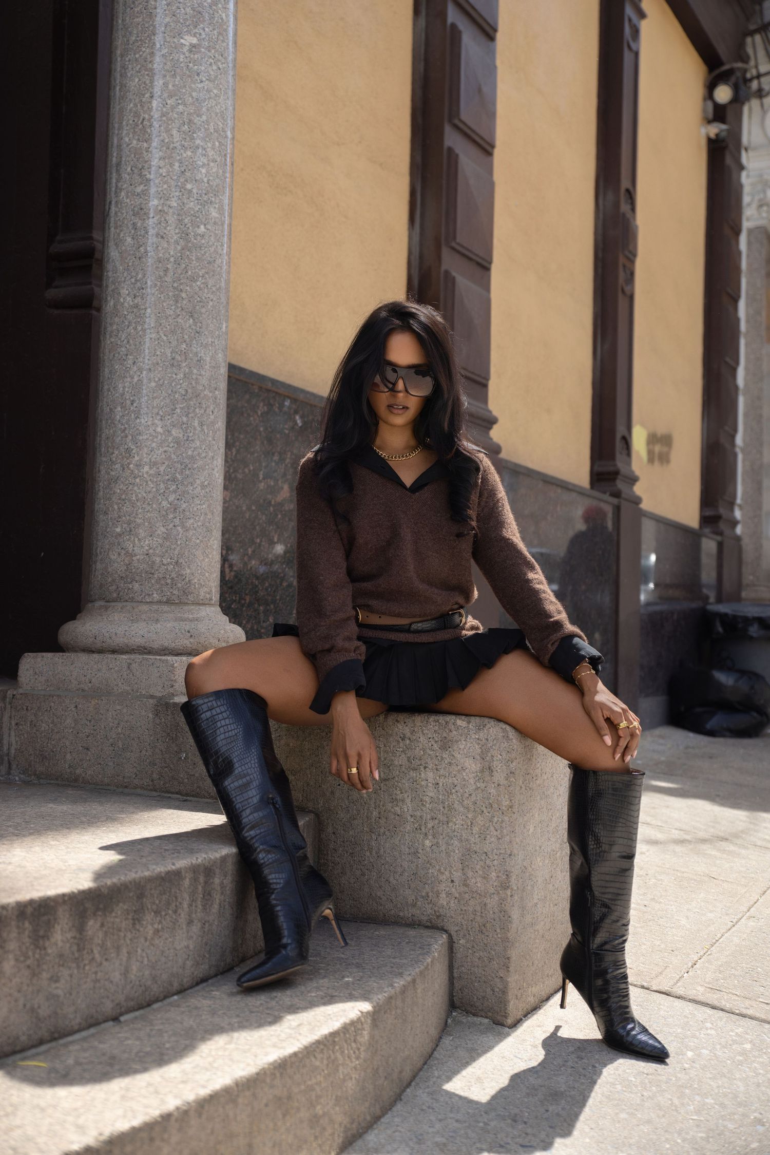 Woman in brown sweater and black mini skirt with tall boots sits on stone steps.