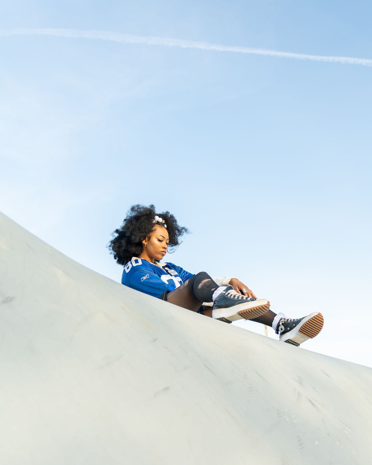 Young woman with curly hair sitting on skate ramp slope under clear blue sky.