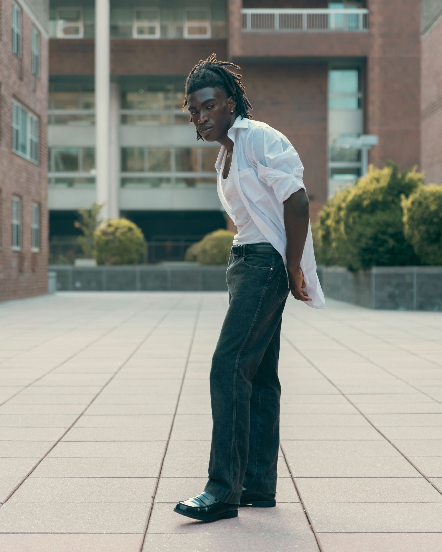 Young Black man with dreadlocks in white shirt and dark jeans posing on urban plaza tiles.