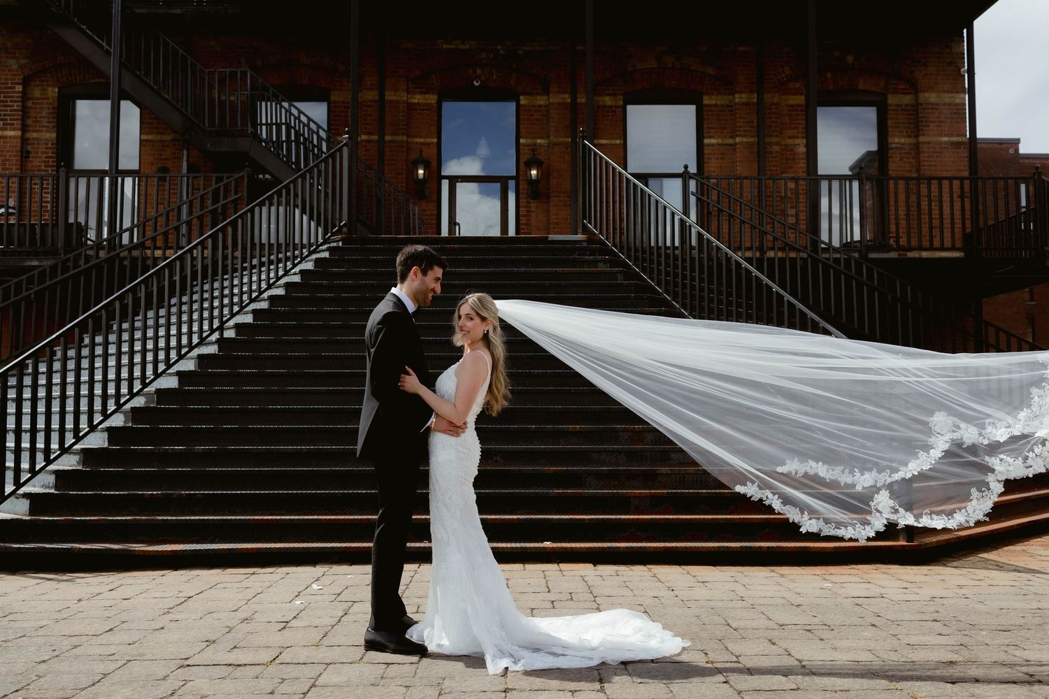 A long flowing white wedding veil billows in the wind against brick architecture and steps.