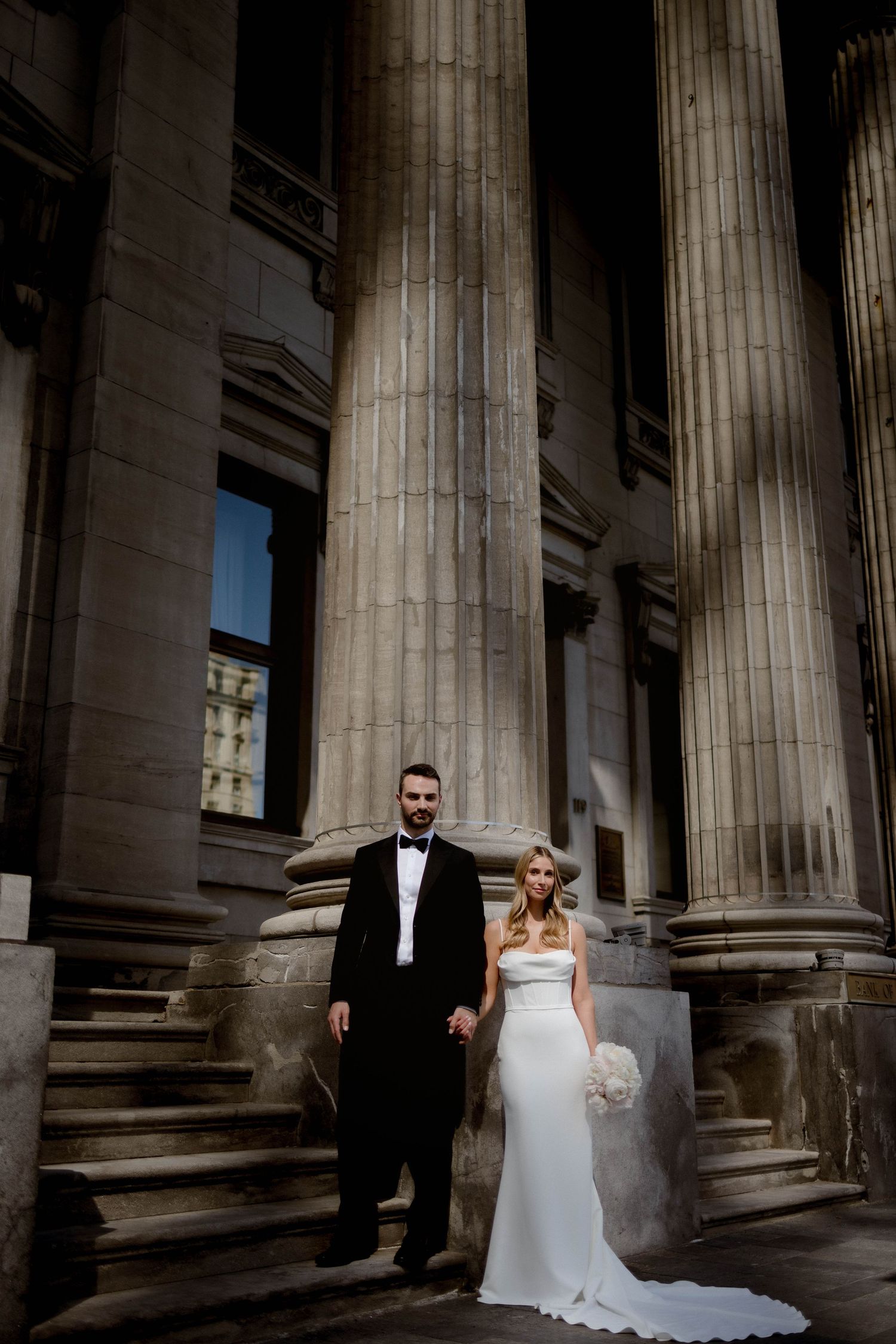 A wedding couple poses elegantly between towering classical stone columns at a grand building entrance.
