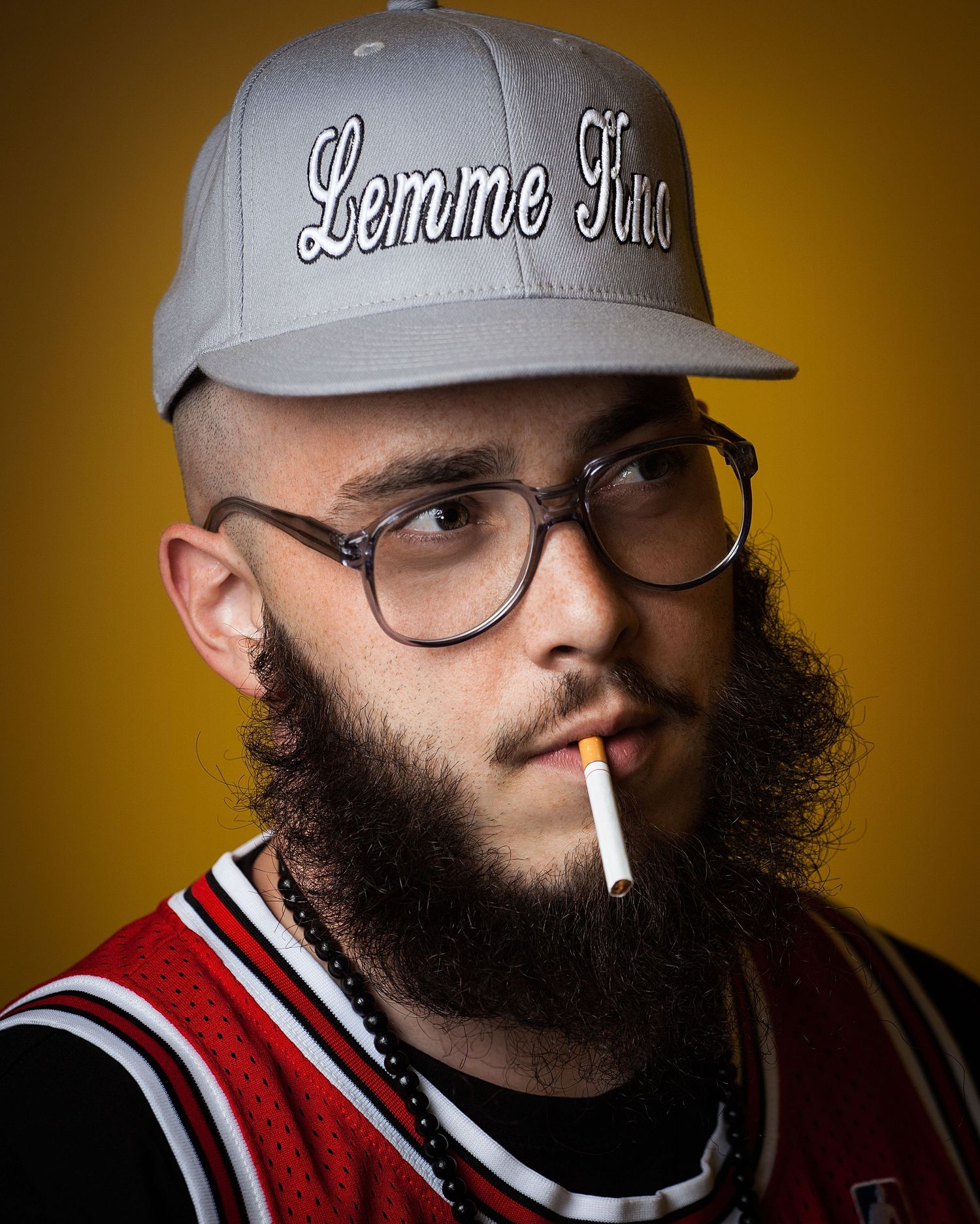 Portrait of a bearded person wearing glasses and a snapback cap with an unlit cigarette.