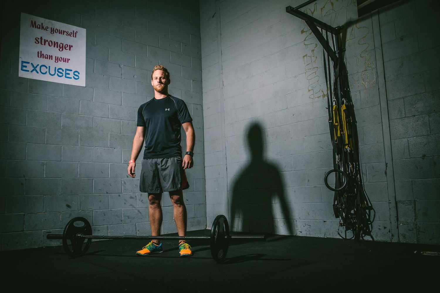 An athlete stands in a dimly lit gym with exercise equipment and his shadow visible on the wall.