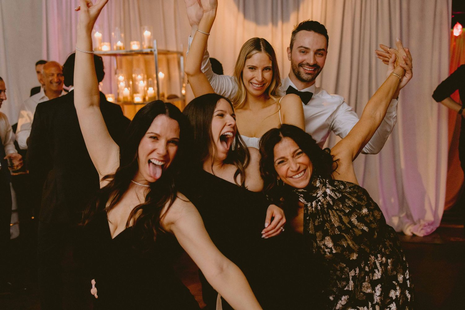 A celebratory group poses exuberantly at a wedding reception with festive lighting in the background.
