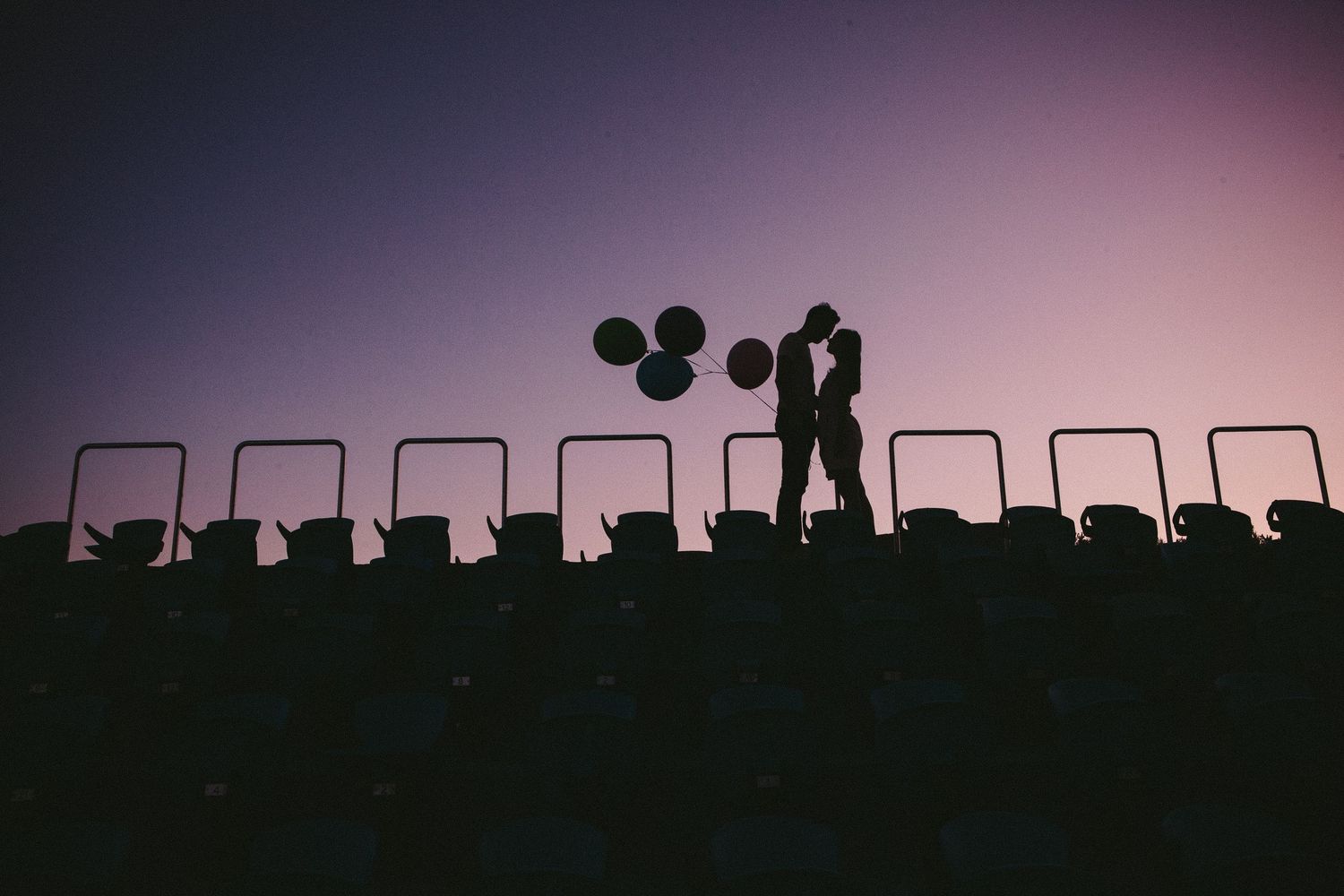 Couple embracing against sunset sky with balloon silhouettes and fence railings.