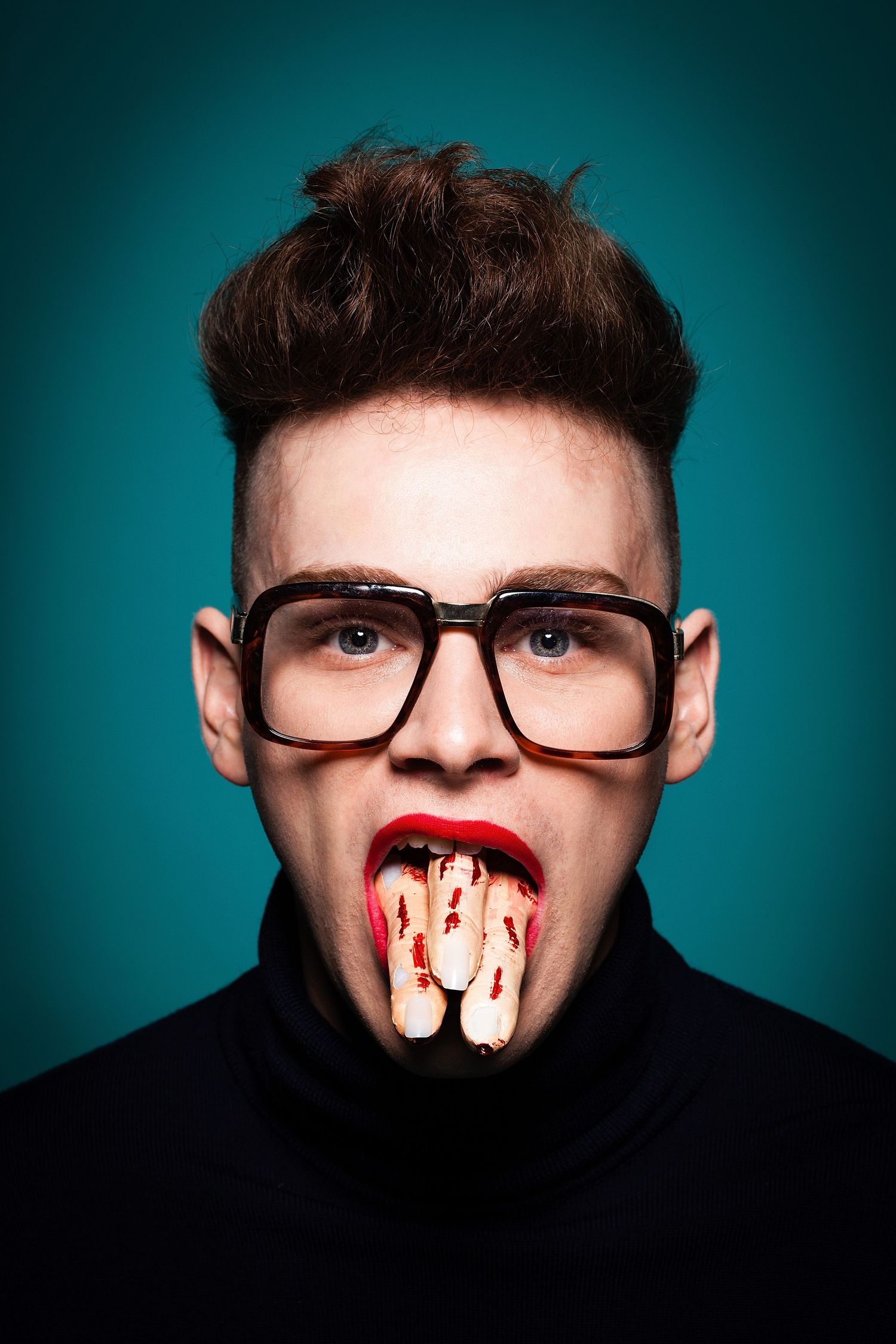 Creative portrait of a person in glasses with theatrical bloody makeup in their mouth against teal background.