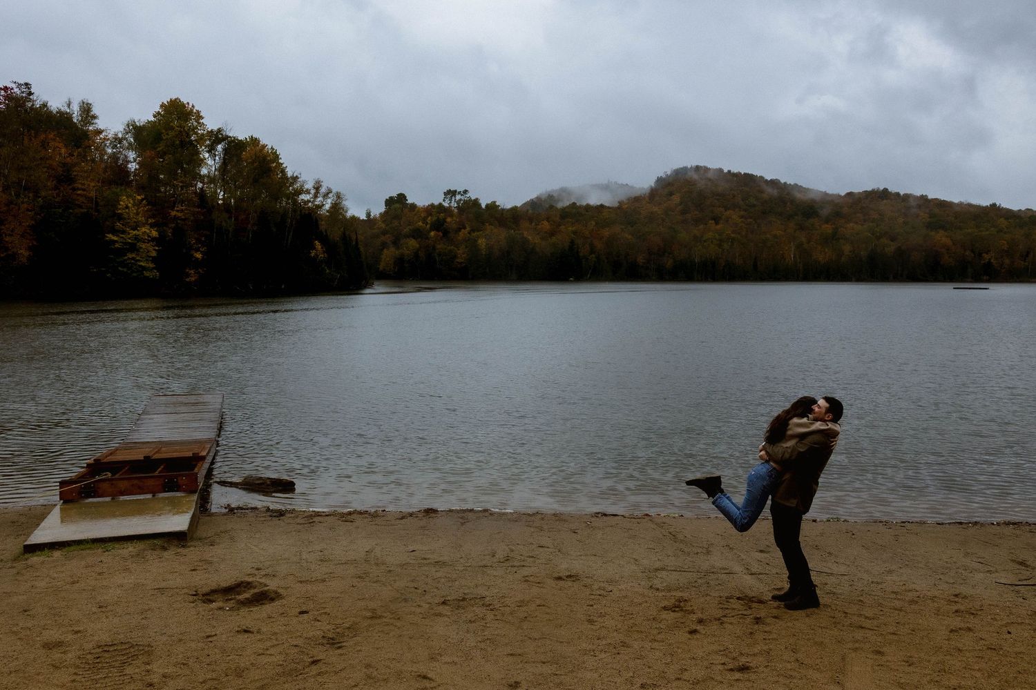 Two people share a romantic moment on a lake beach with cloudy skies and autumn trees.