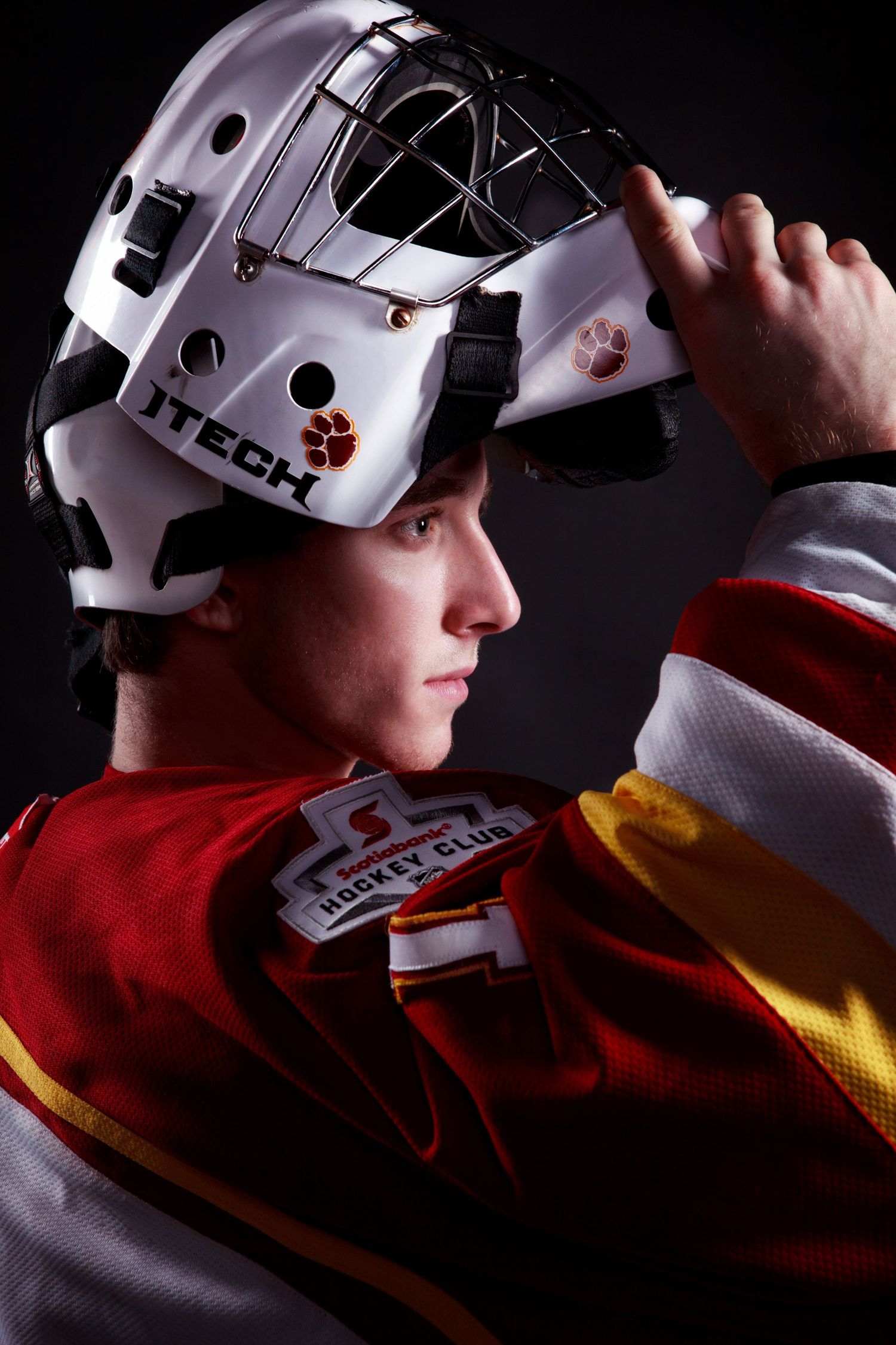 Hockey goalie in white mask and red jersey shown in dramatic side profile.
