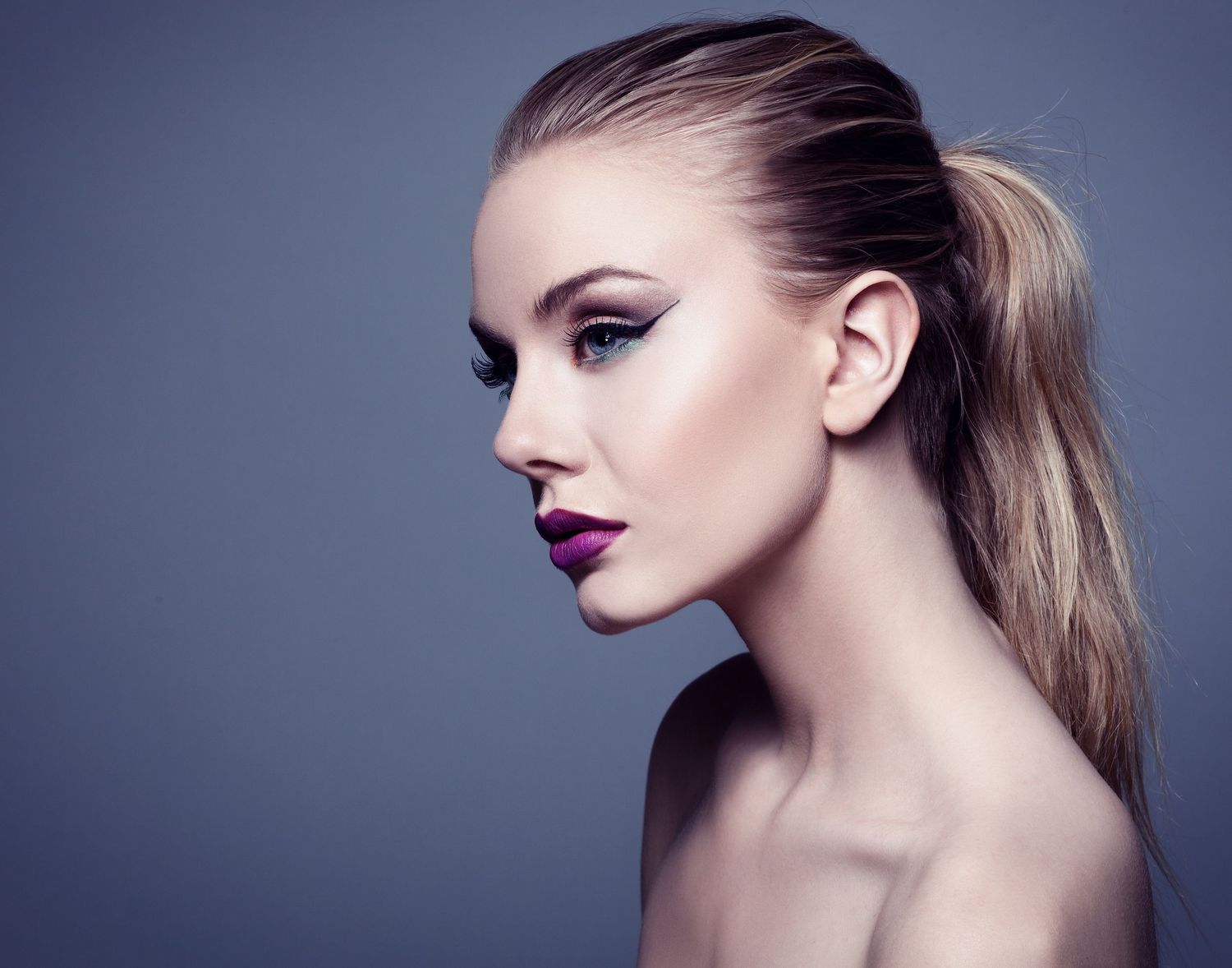 Professional beauty portrait with dramatic makeup and slicked back blonde hair against dark background.