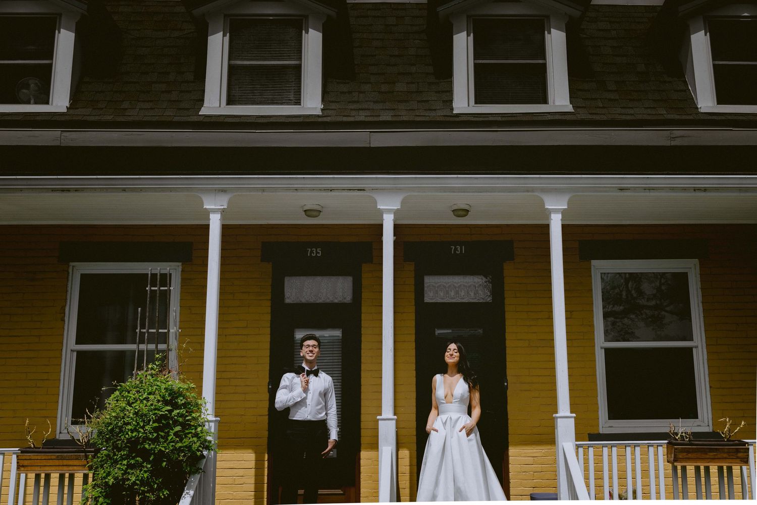 A couple in wedding attire poses in front of a charming yellow house with a traditional porch.