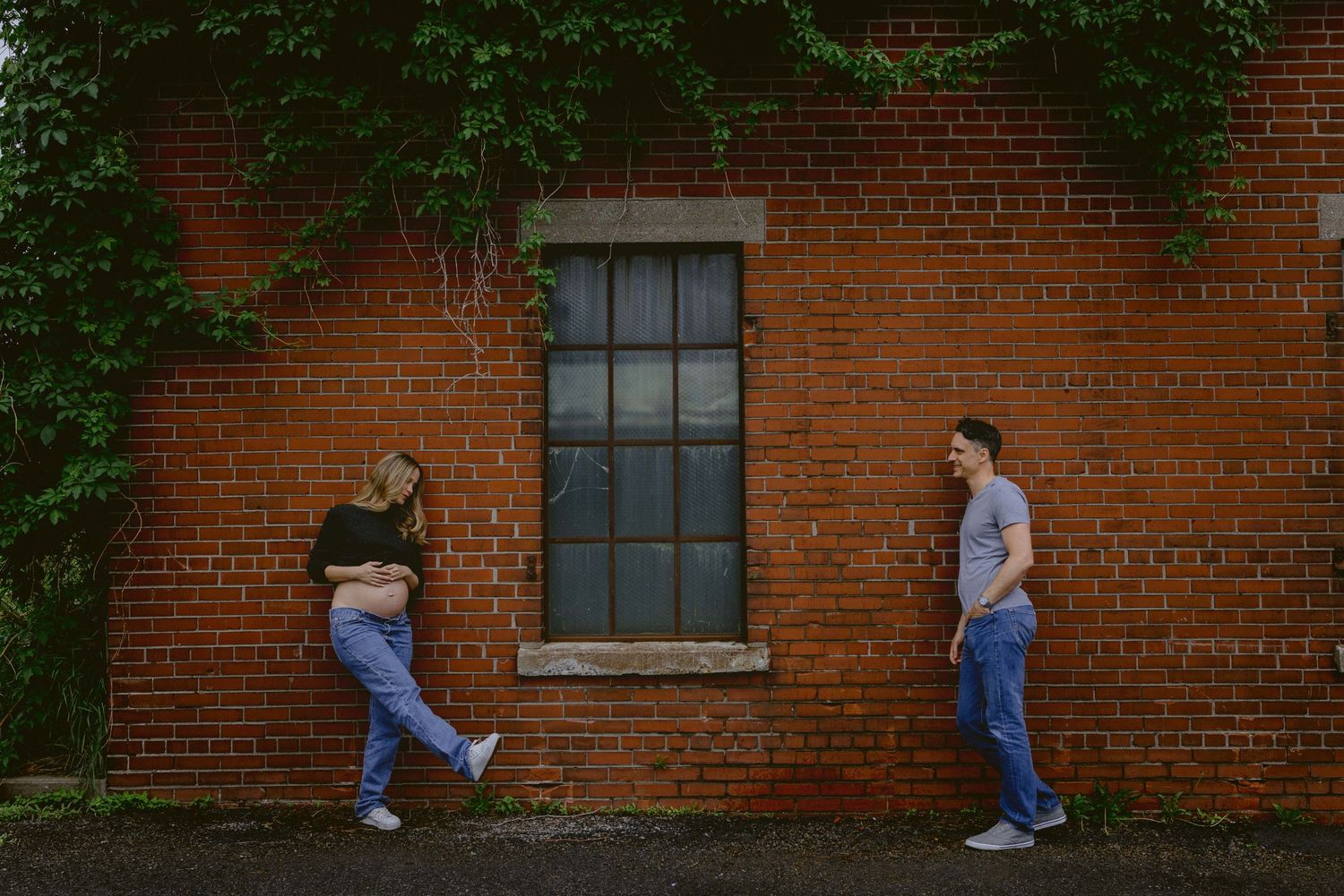 Two people pose playfully against a rustic brick wall with a large window between them.