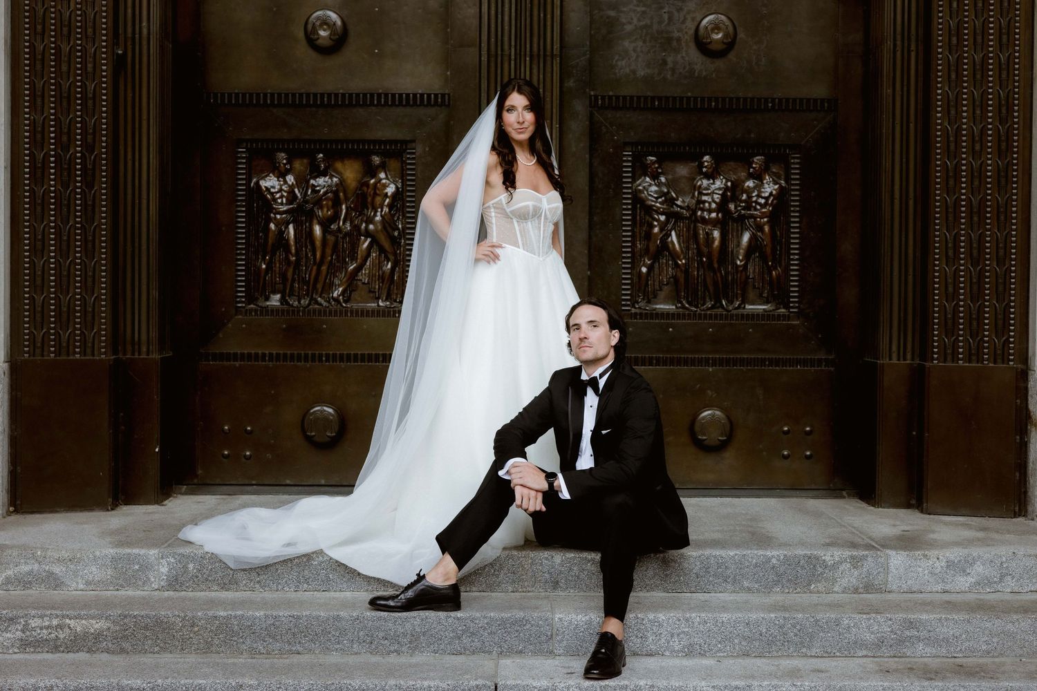 A couple poses dramatically on stone steps, with the bride standing tall in a flowing veil while the groom sits below.