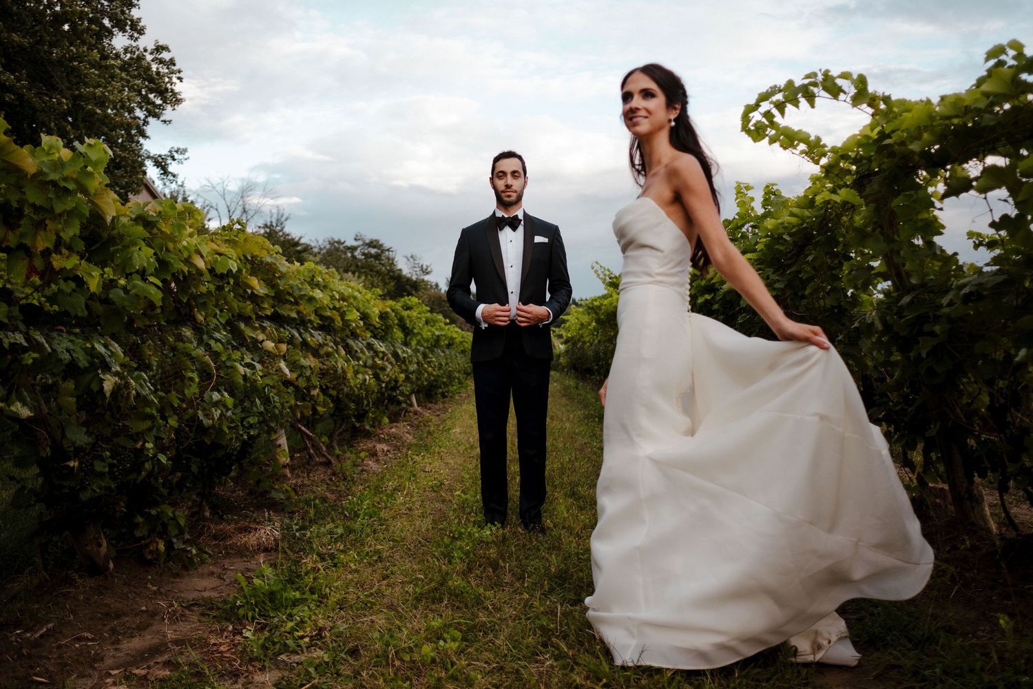 Bride in flowing white dress poses with groom in vineyard during sunset photo session.