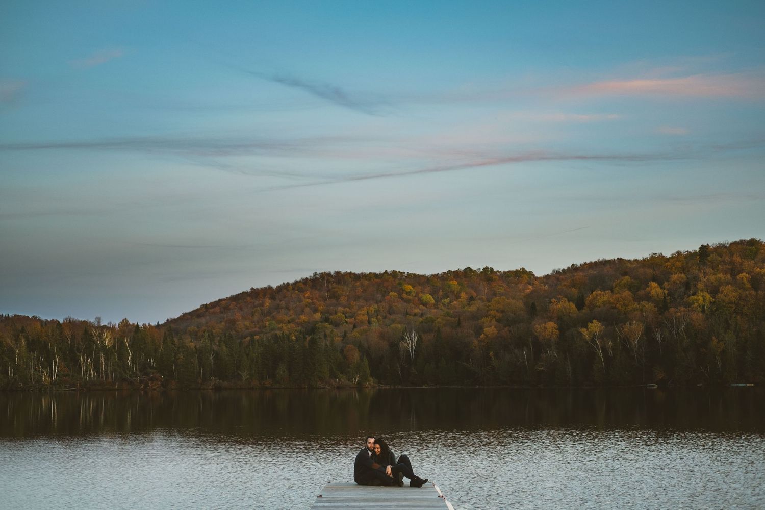 Couple sitting together on dock overlooking autumn lake at sunset.