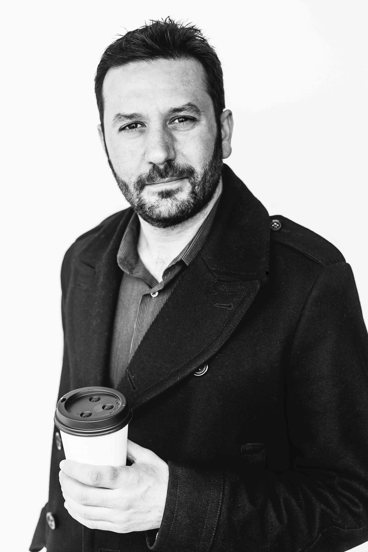 Professional headshot of man in black coat holding coffee cup against white background.
