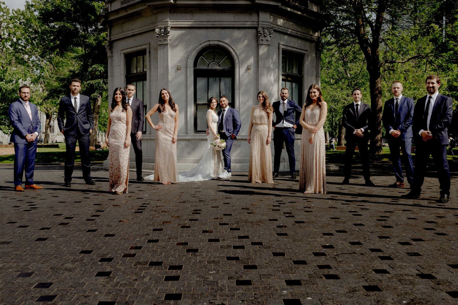 Wedding party members lined up on cobblestone pavement surrounded by lush greenery.