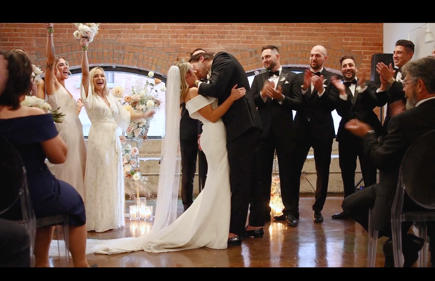 Wedding couple shares first kiss surrounded by wedding party in elegant indoor ceremony space.
