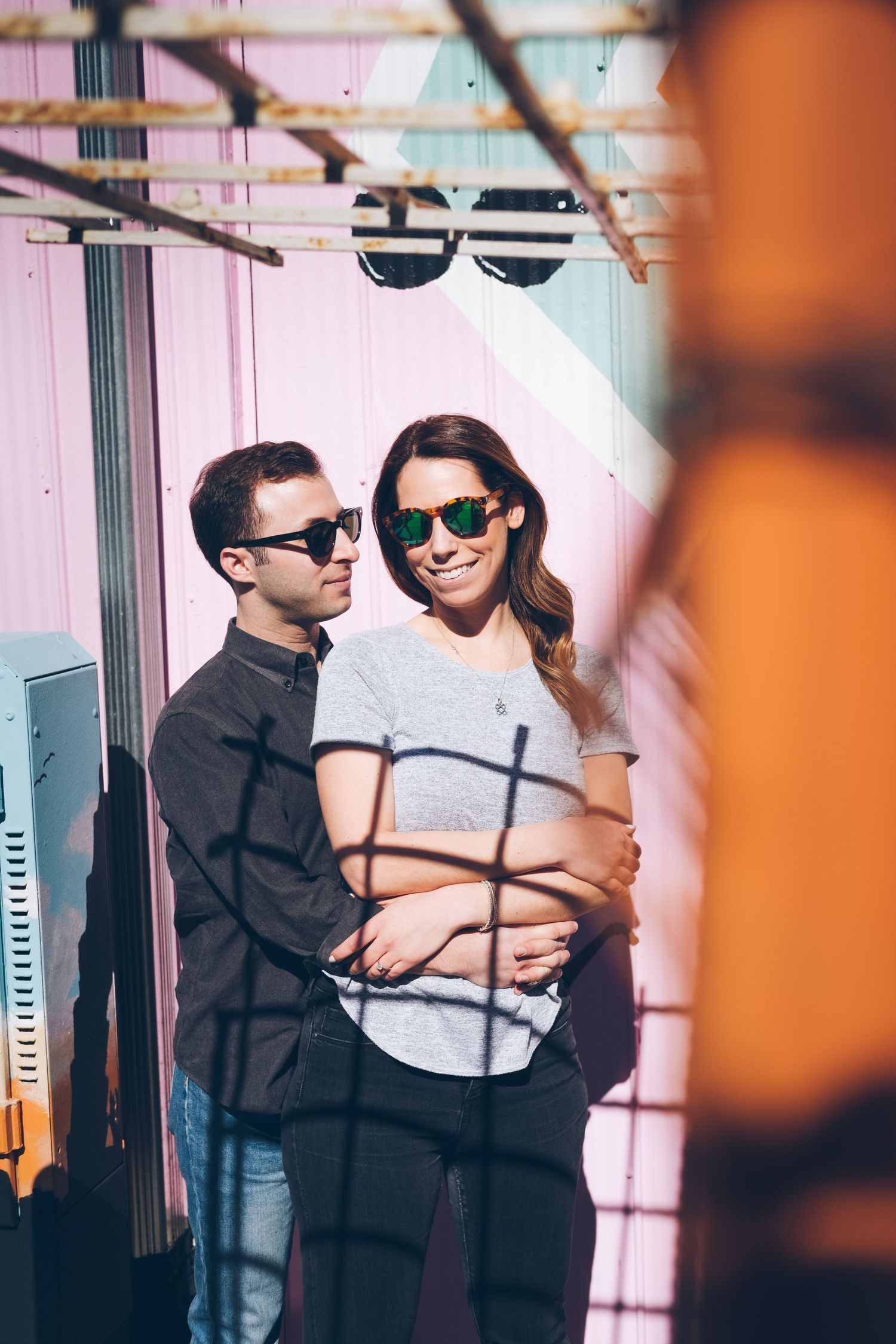 Stylish couple in sunglasses posing against pink wall with geometric shadows.