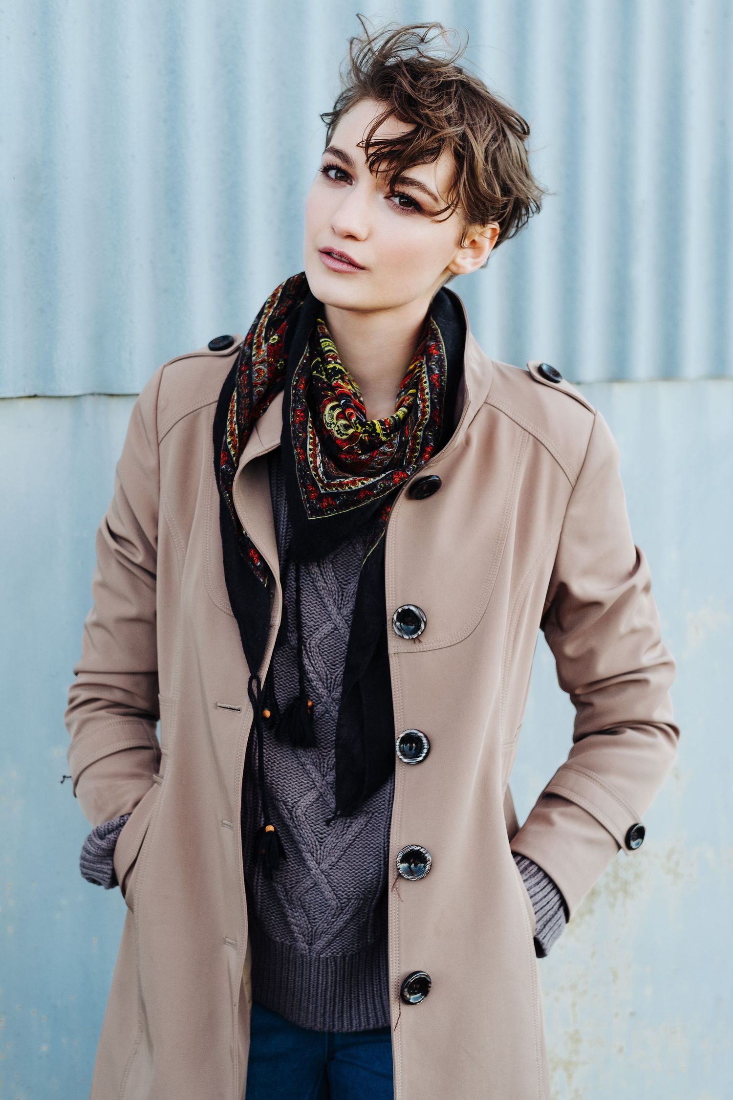 Fashion editorial of model in beige coat and dark sweater against blue background.