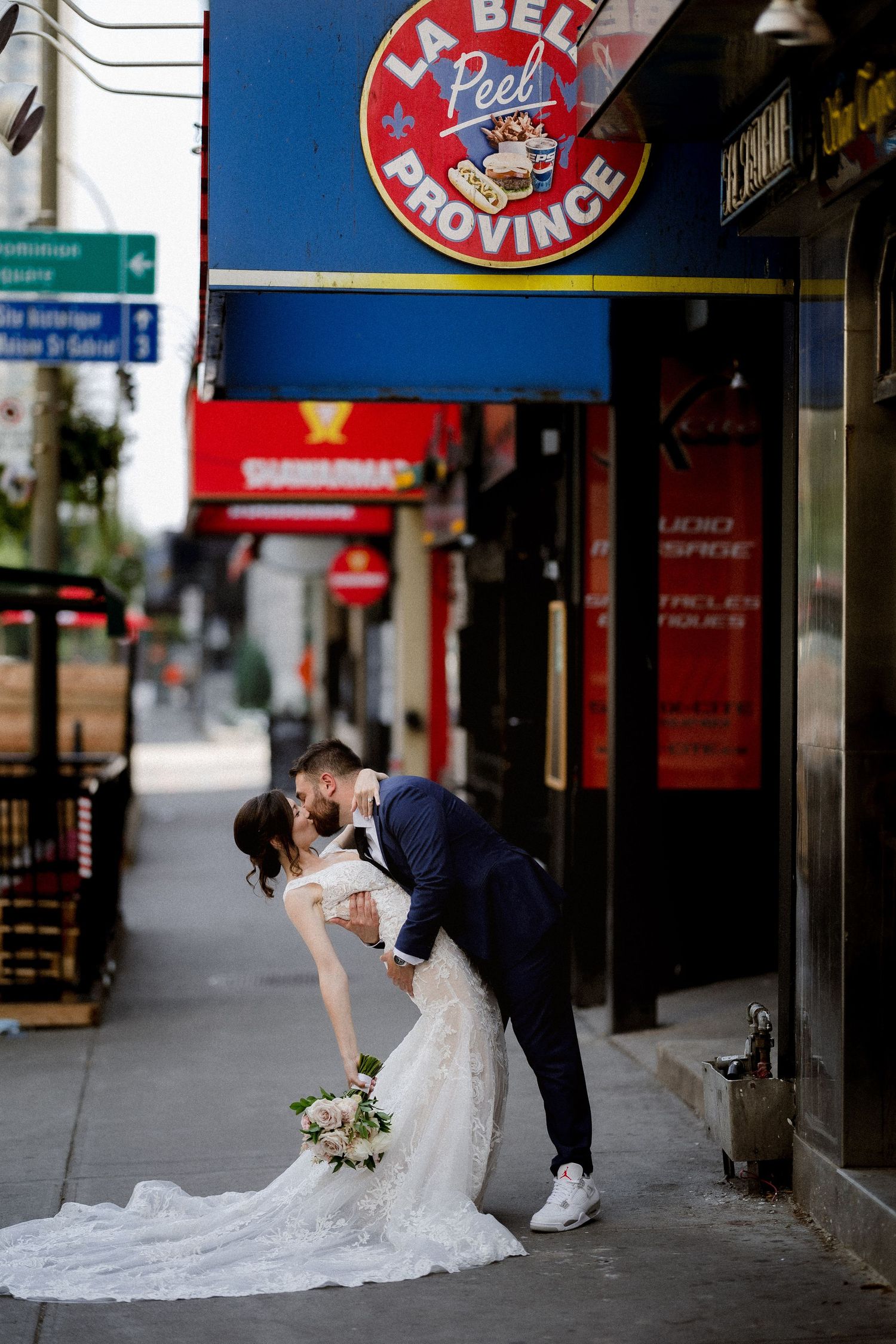 A couple in wedding attire poses in front of storefront signs along an urban street.