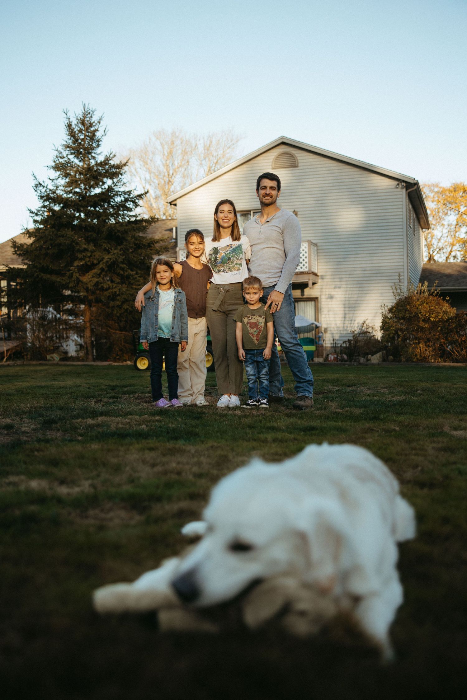 Family stands on their lawn with a white house in background while their dog rests in foreground.