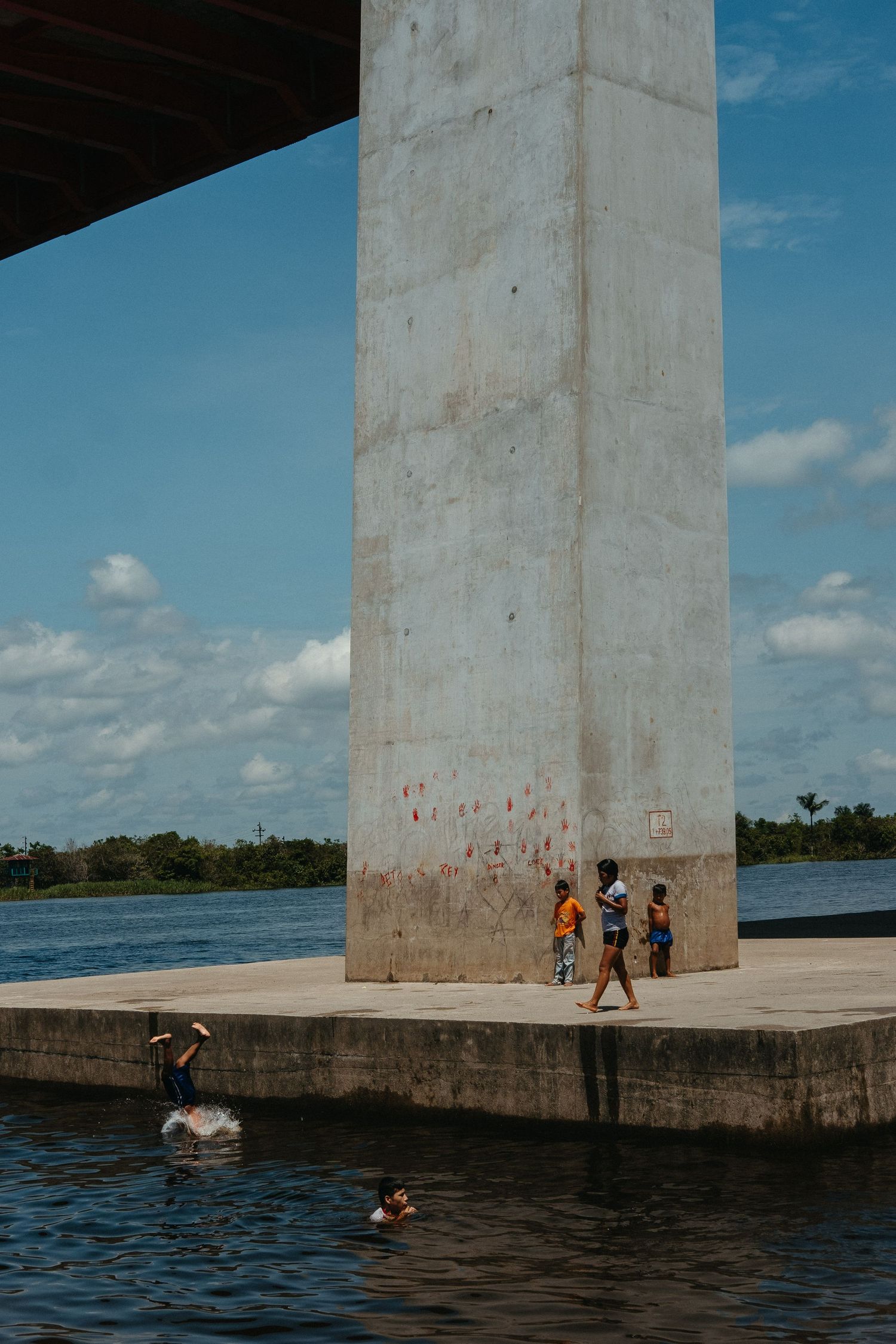 People swimming and playing near large concrete bridge pillar over blue water.