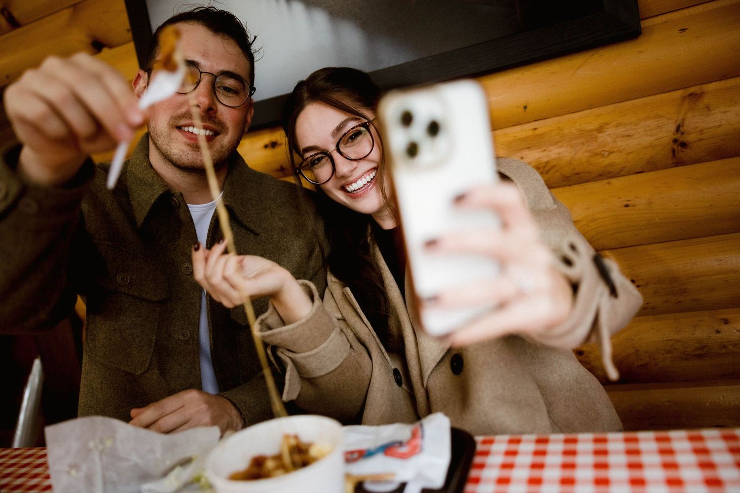 Couple taking selfie while enjoying meal at rustic log cabin restaurant with checkered tablecloth.