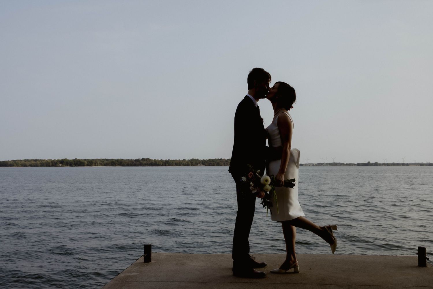 A couple shares an intimate waterfront moment at sunset with a dramatic cloudy sky backdrop.