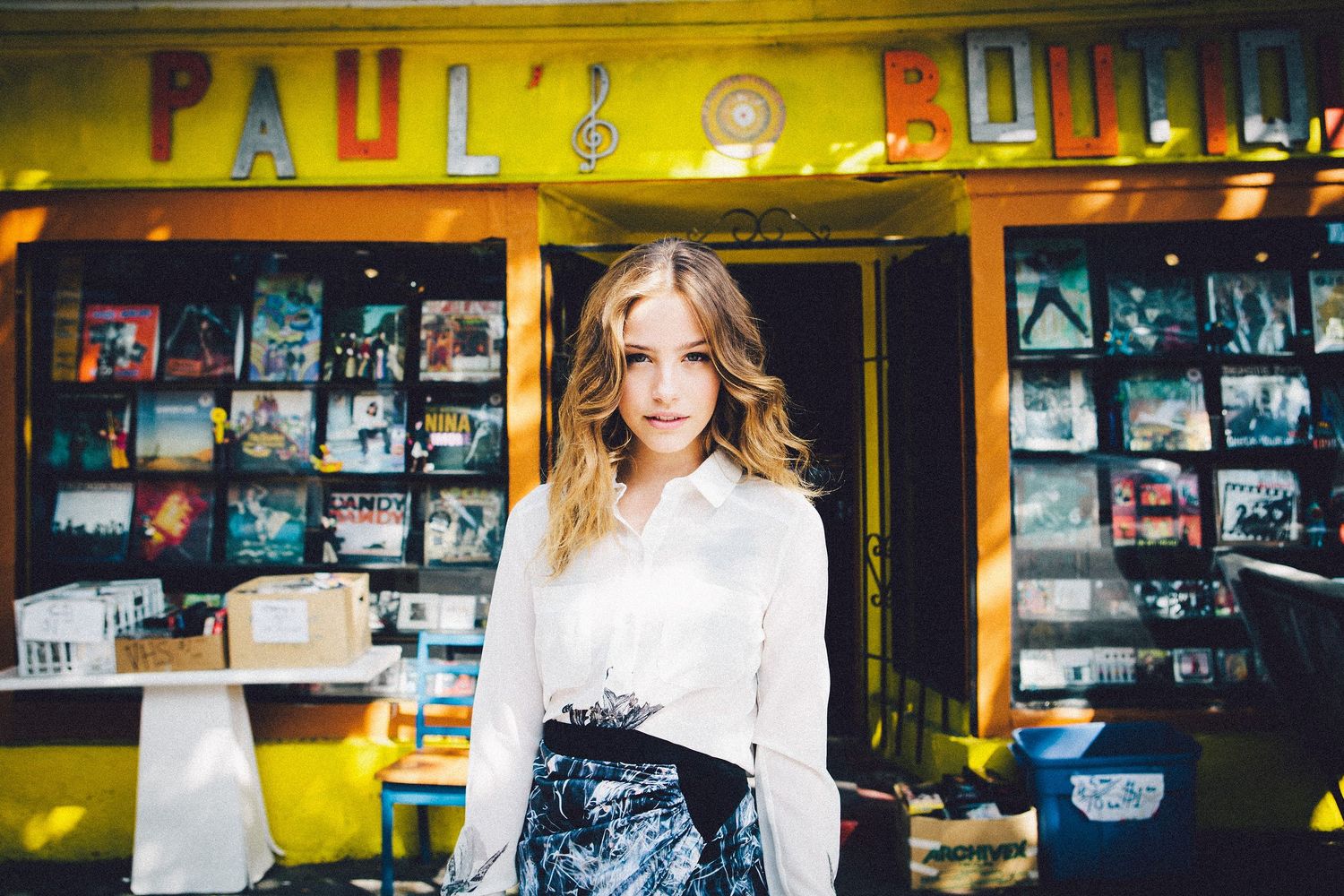 Woman in white top poses in front of colorful record store entrance with vintage signage.