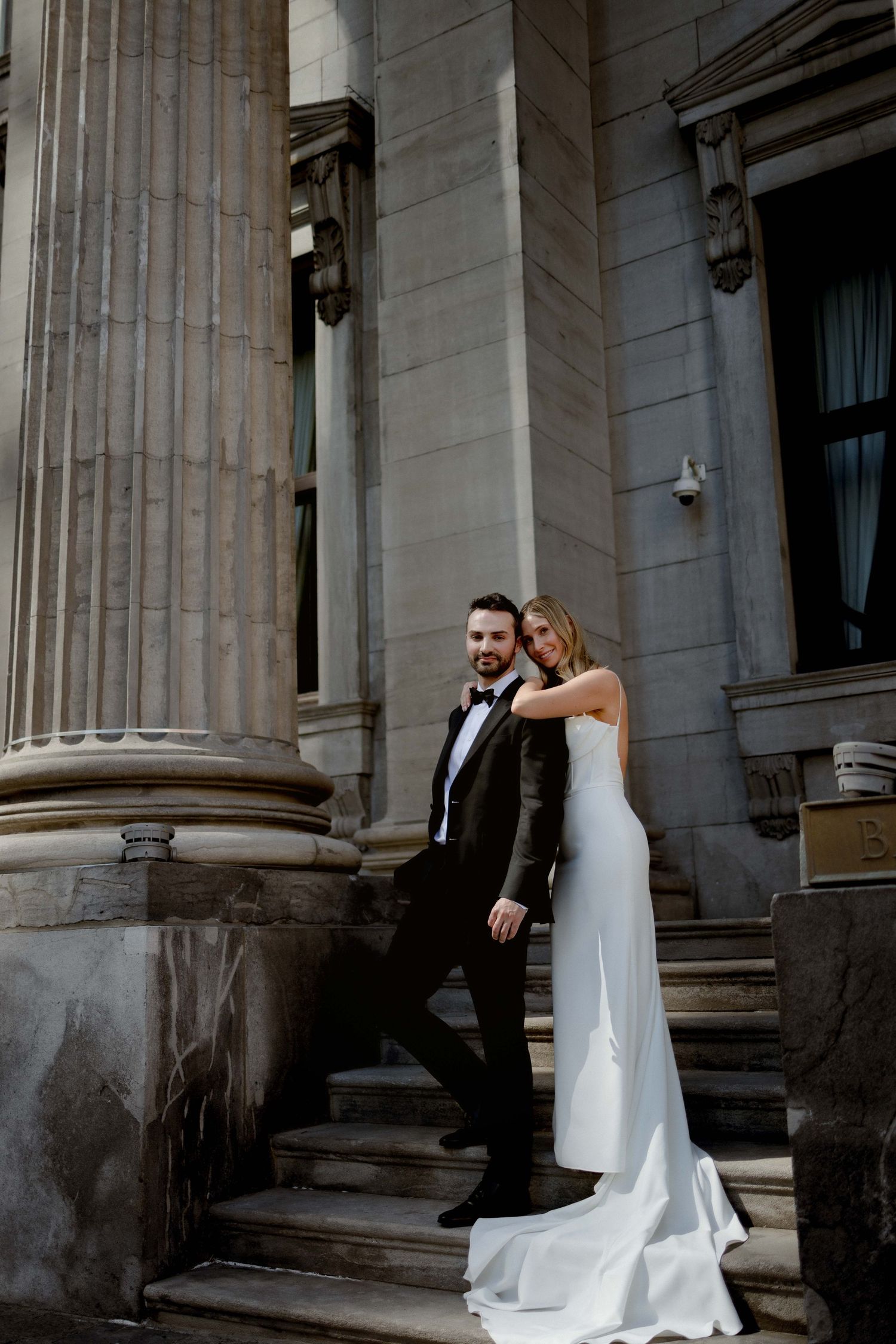 A couple embraces on stone steps between elegant classical columns in formal wedding attire.
