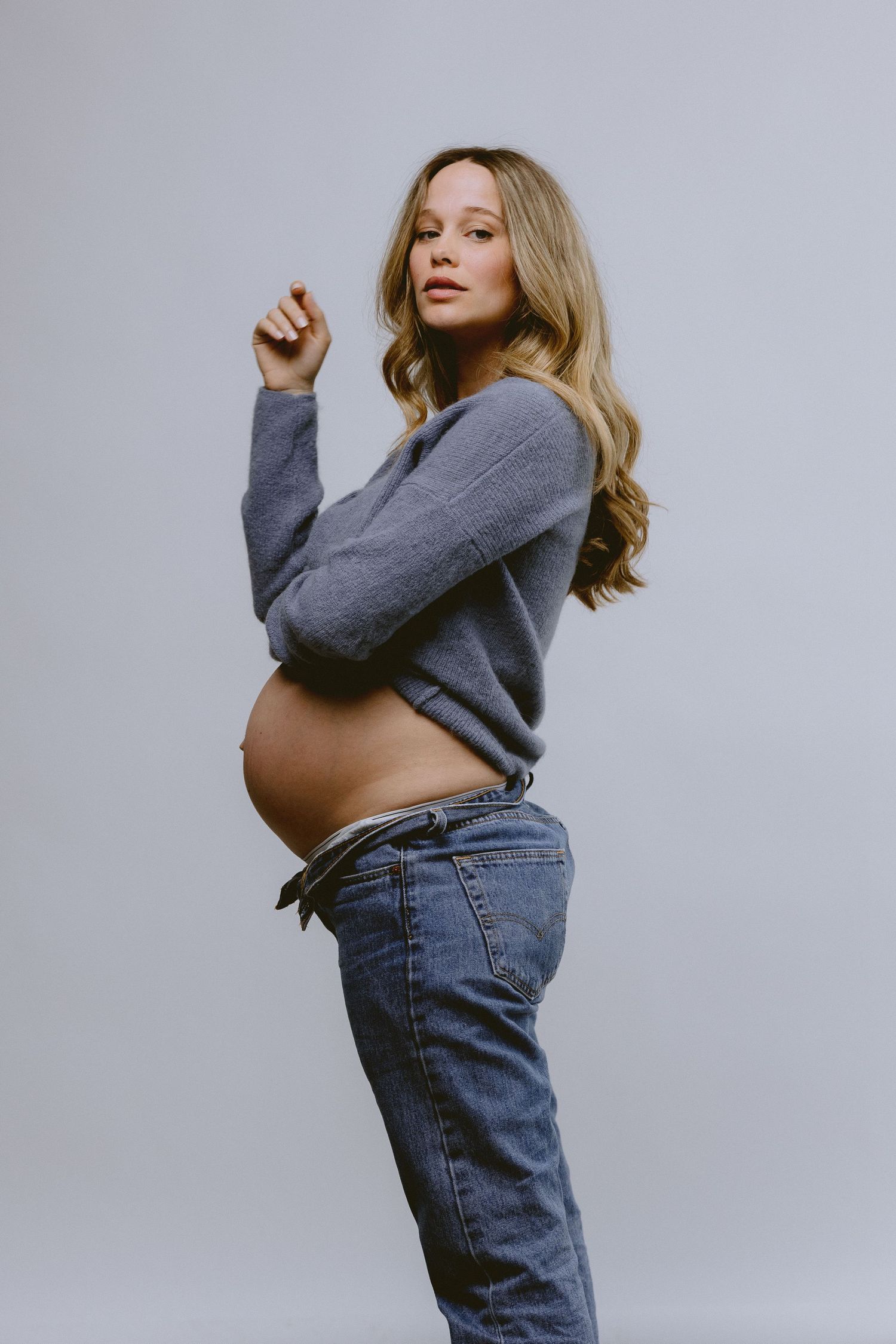 Side view of pregnant woman in gray sweater showing baby bump against neutral background.