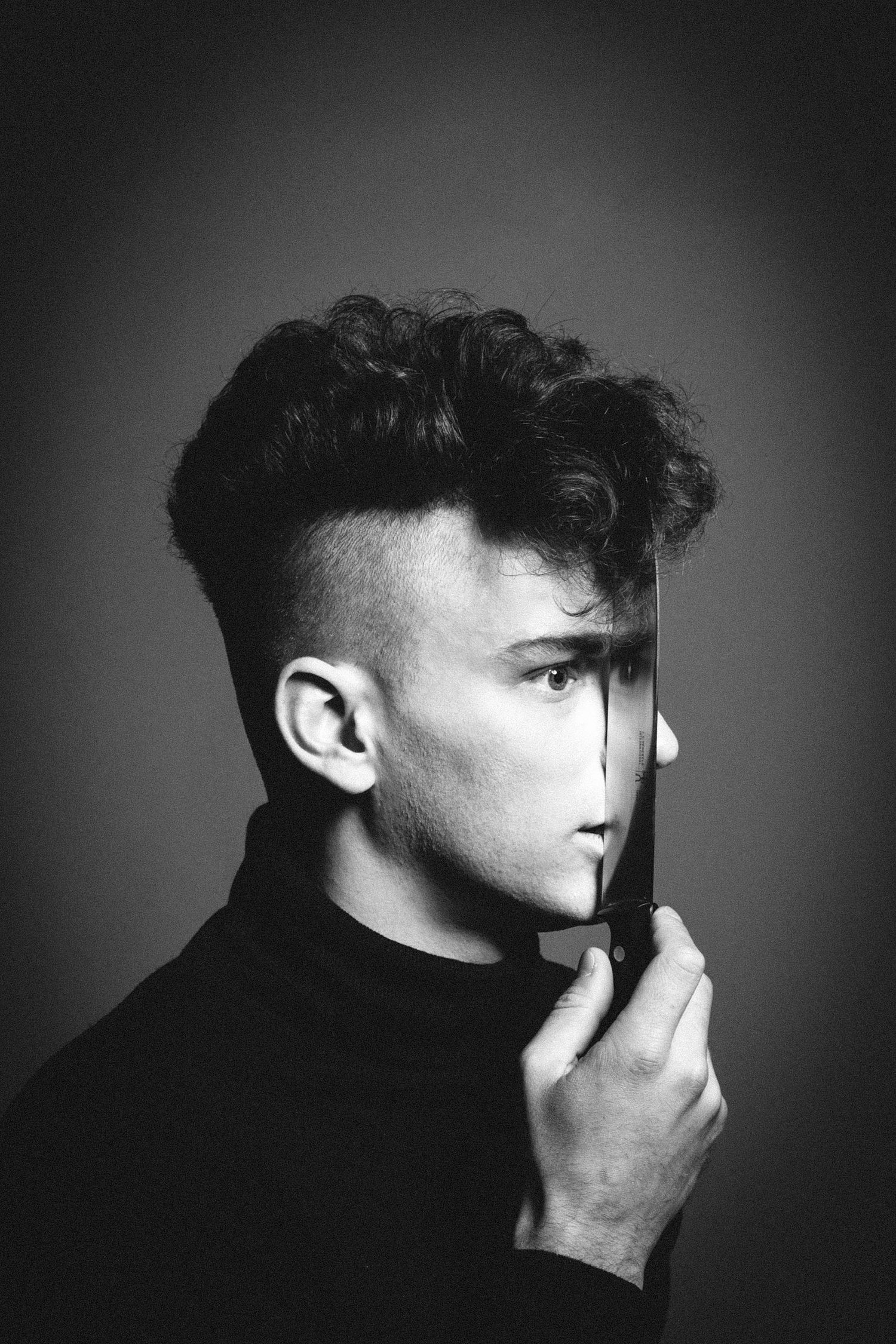 Dramatic black and white portrait of a person with an undercut hairstyle holding a reflective object near their face.