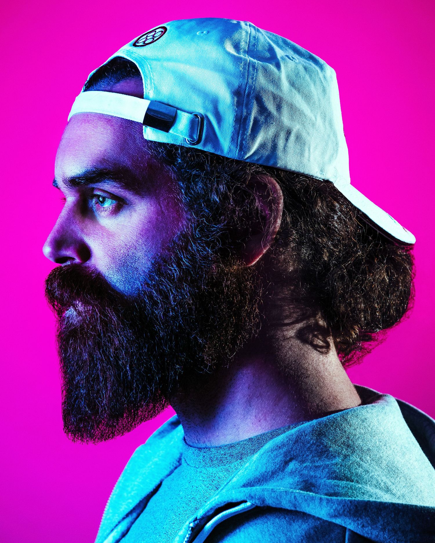 Profile portrait of a bearded person wearing a blue baseball cap against a vibrant pink background.