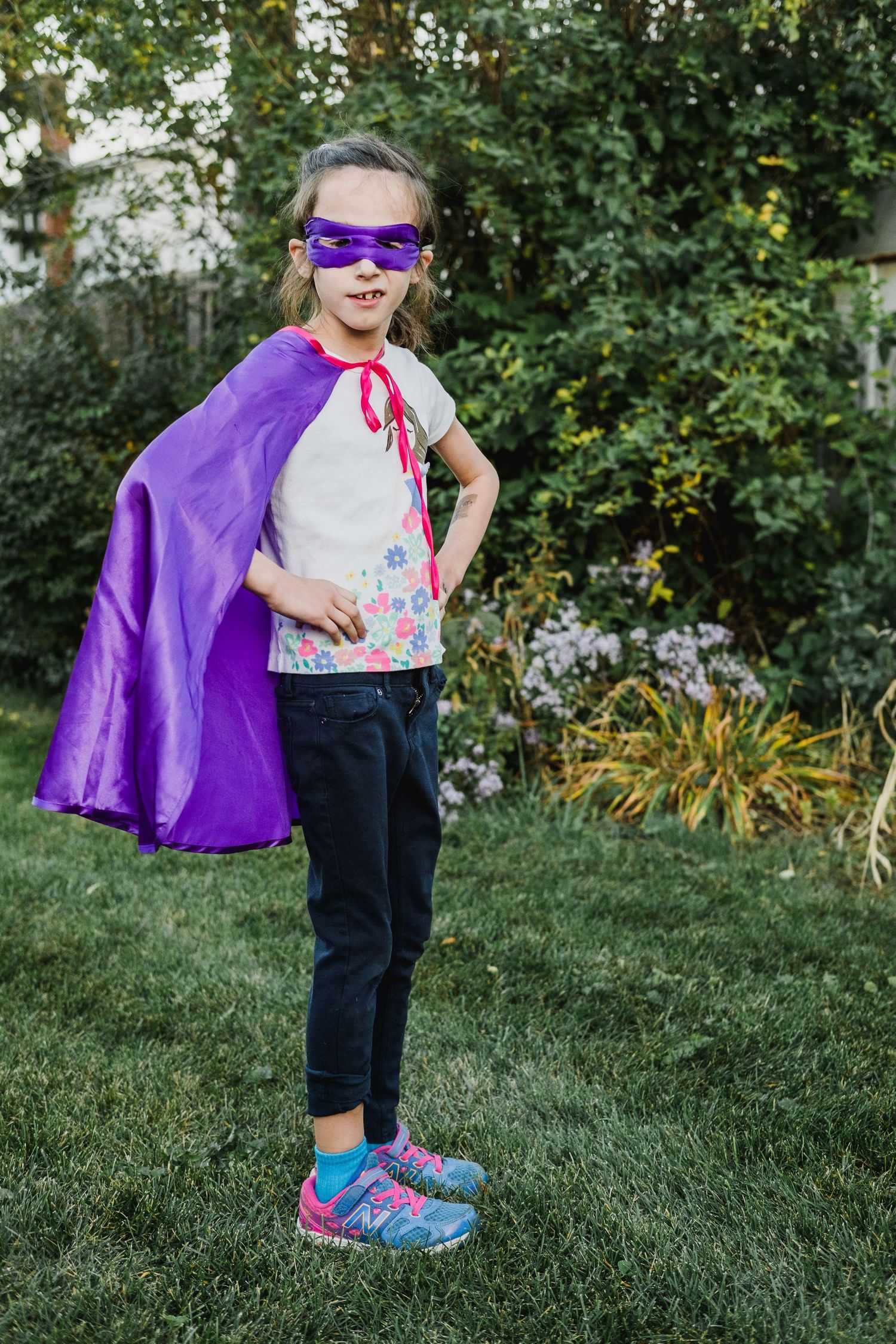 Young superhero posing in purple cape and mask in backyard garden.