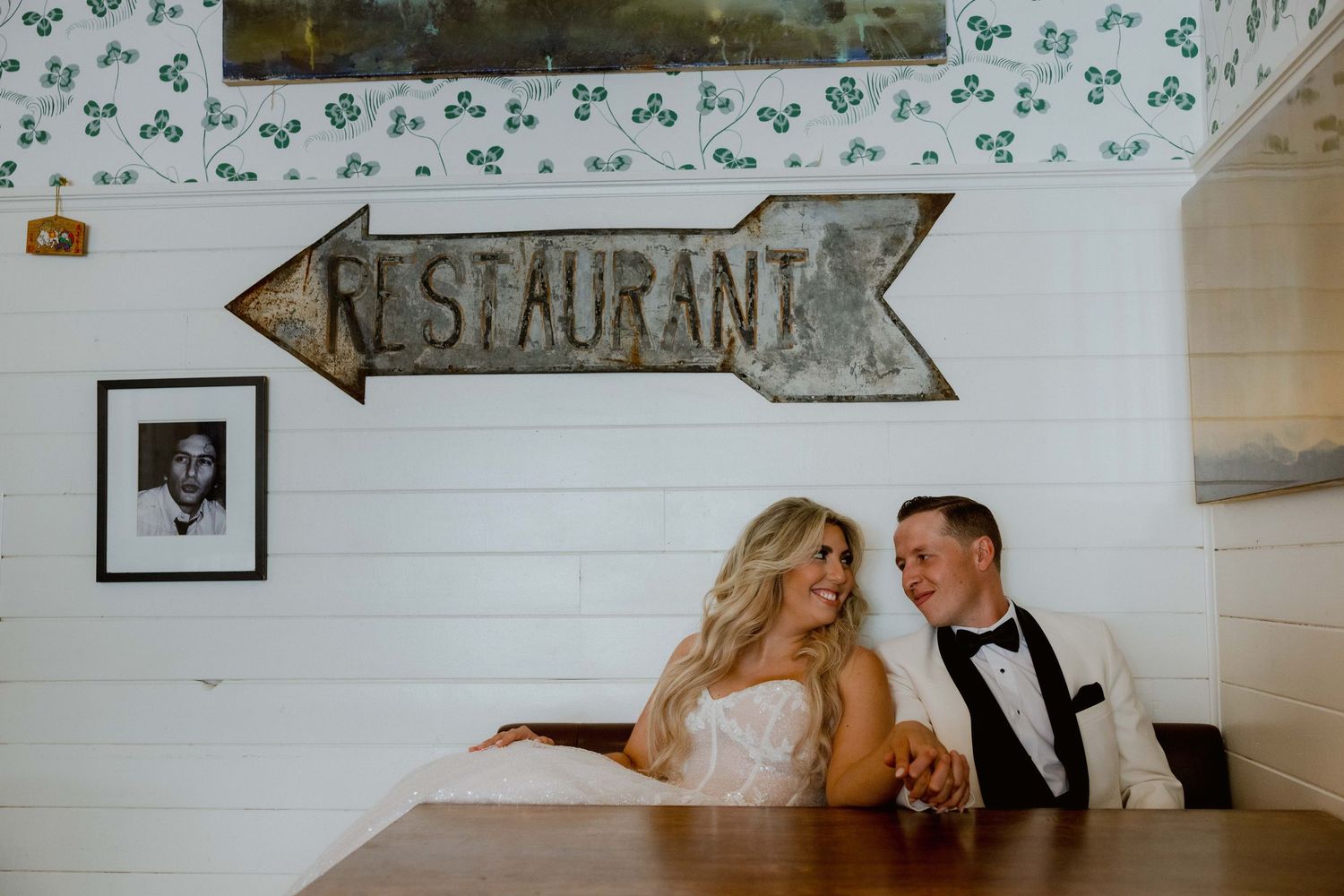 A couple sits at a table beneath a rustic restaurant sign in a charming white-walled setting.