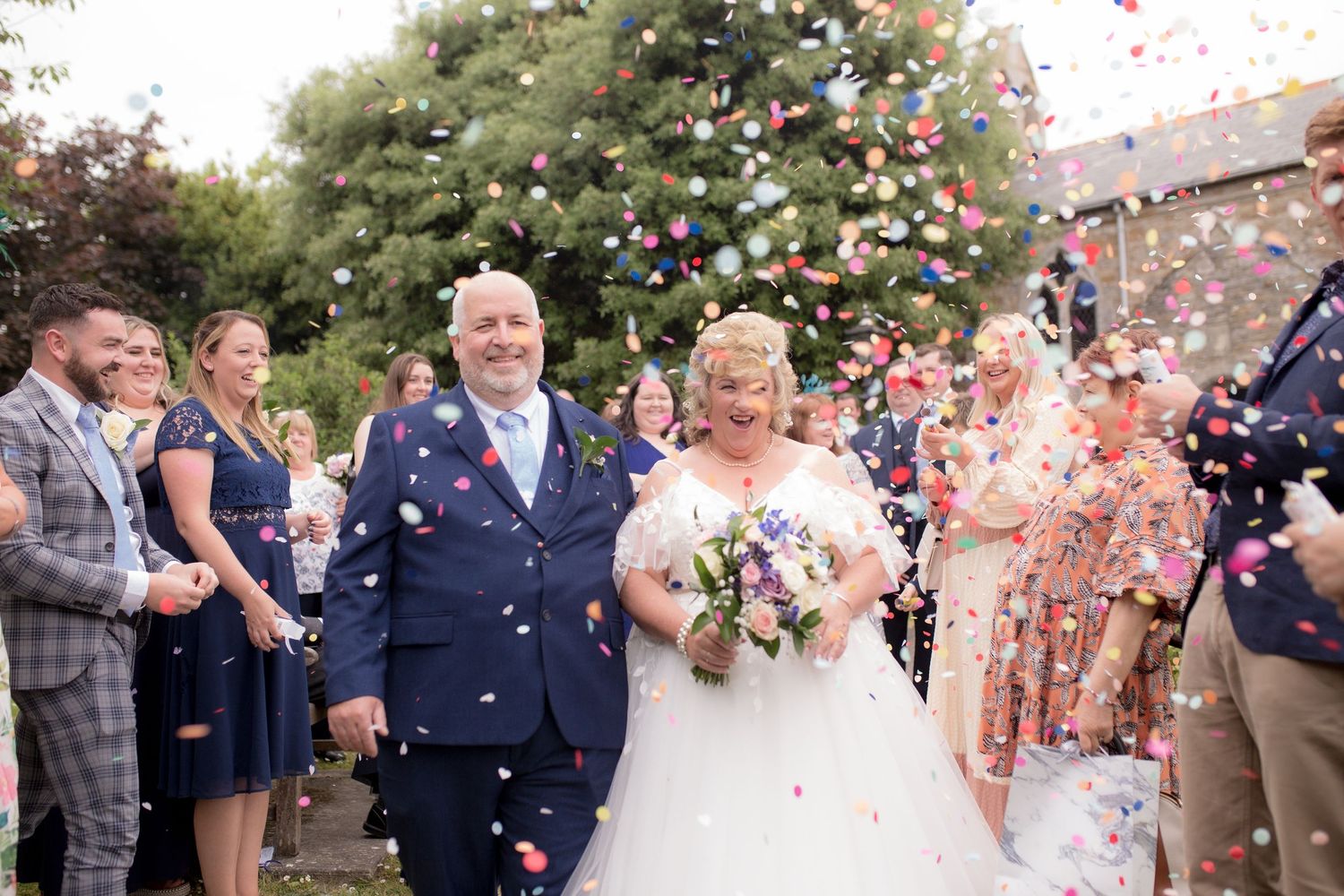 Bride and Groom smile whilst walking through a shower of confetti, surrounded by their loved ones outside the church
