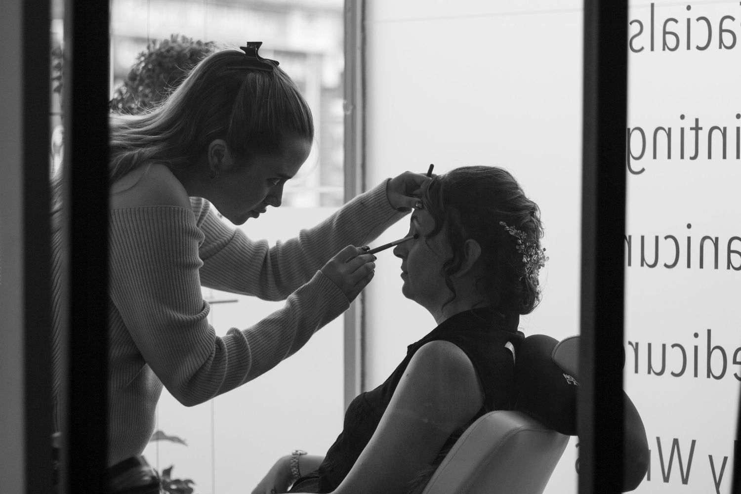 Bride sat having her make up done by a make up artist in a salon. Shot in black and white so they are both silhouetted