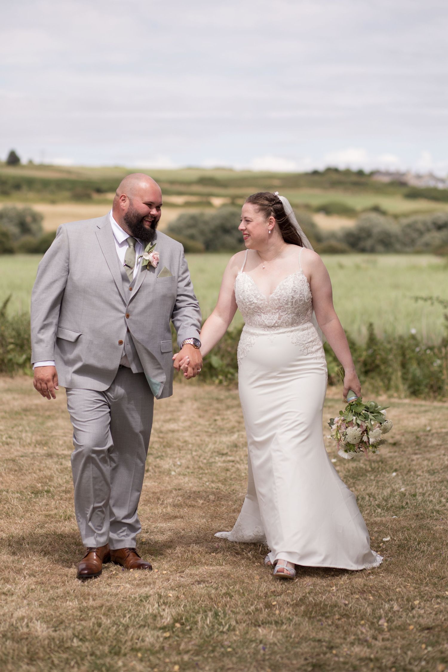 Bride and Groom walk together, eyes facing each other, through green fields in the summer