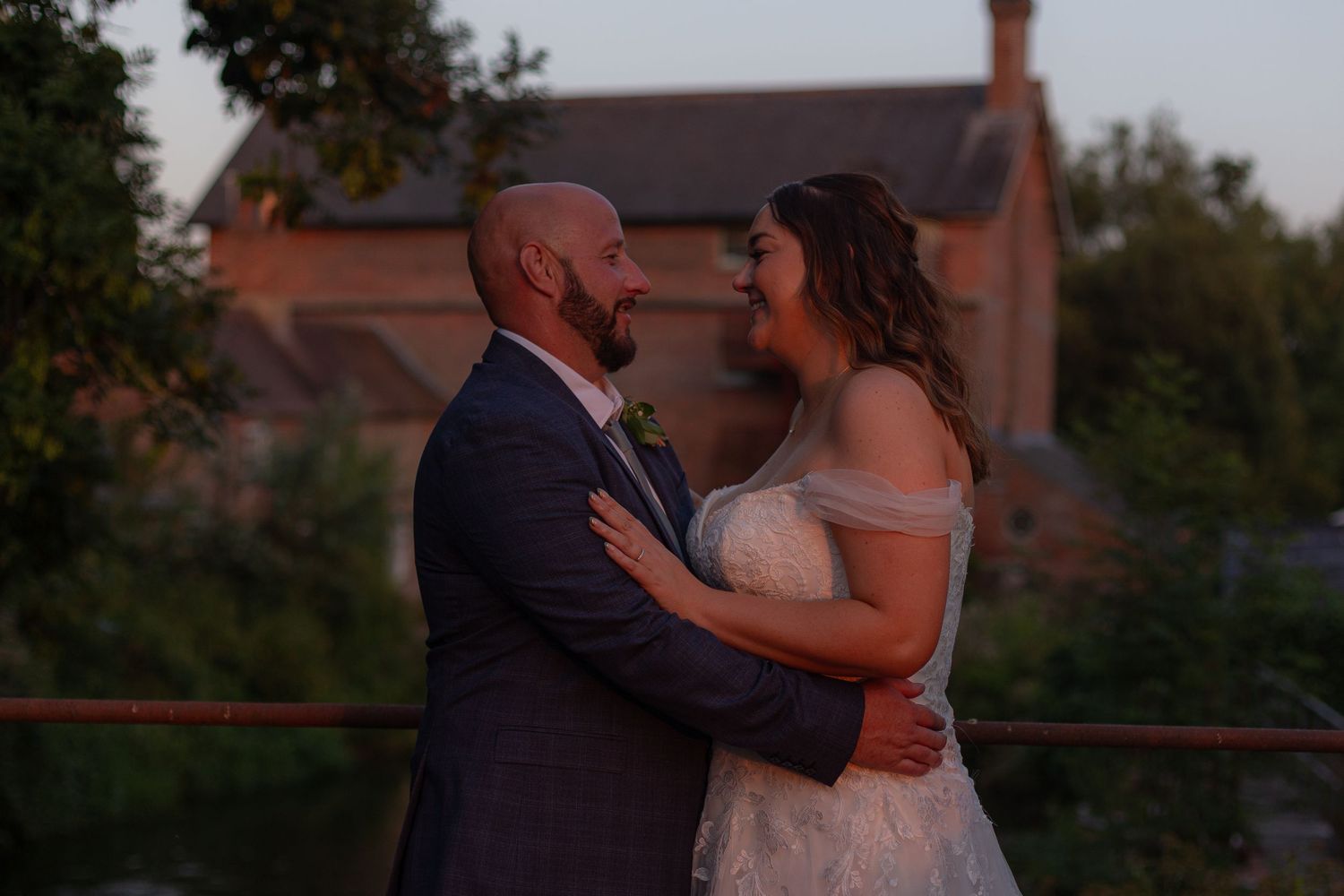 Bride and Groom stand in each others arms, lit by the evening sun and sopley mill standing in the background.