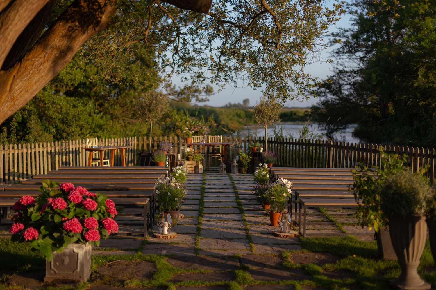 Outdoor ceremony space at Sopley Mill in Dorset. Glowing sunset light, rows of benches and flower pots place under the shade of a tree.
