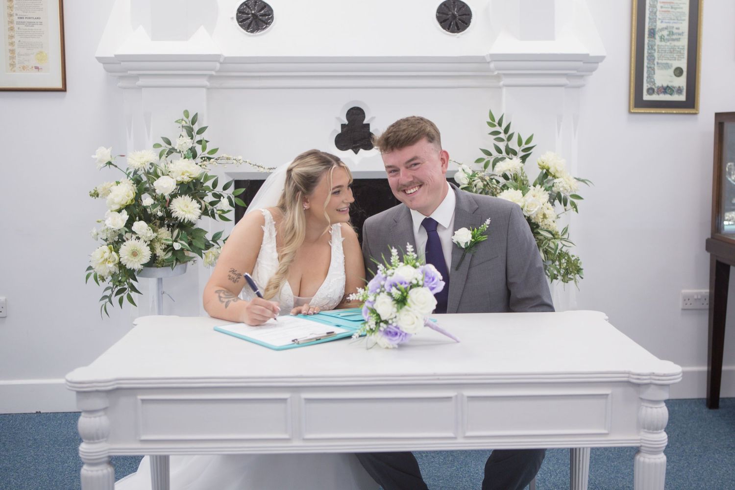 Bride and Groom sit at a table signing their marriage license. The Groom smiles at the camera, and the Bride smiles at her groom.