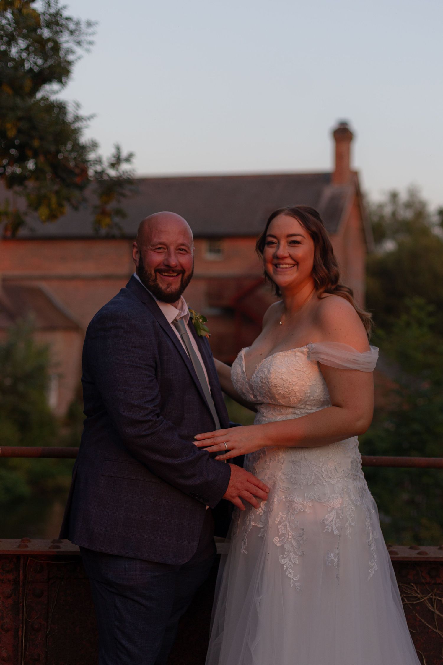 Bride and Groom laugh, looking towards the camera, in the glowing sunset light. Sopley Mill in the background behind them