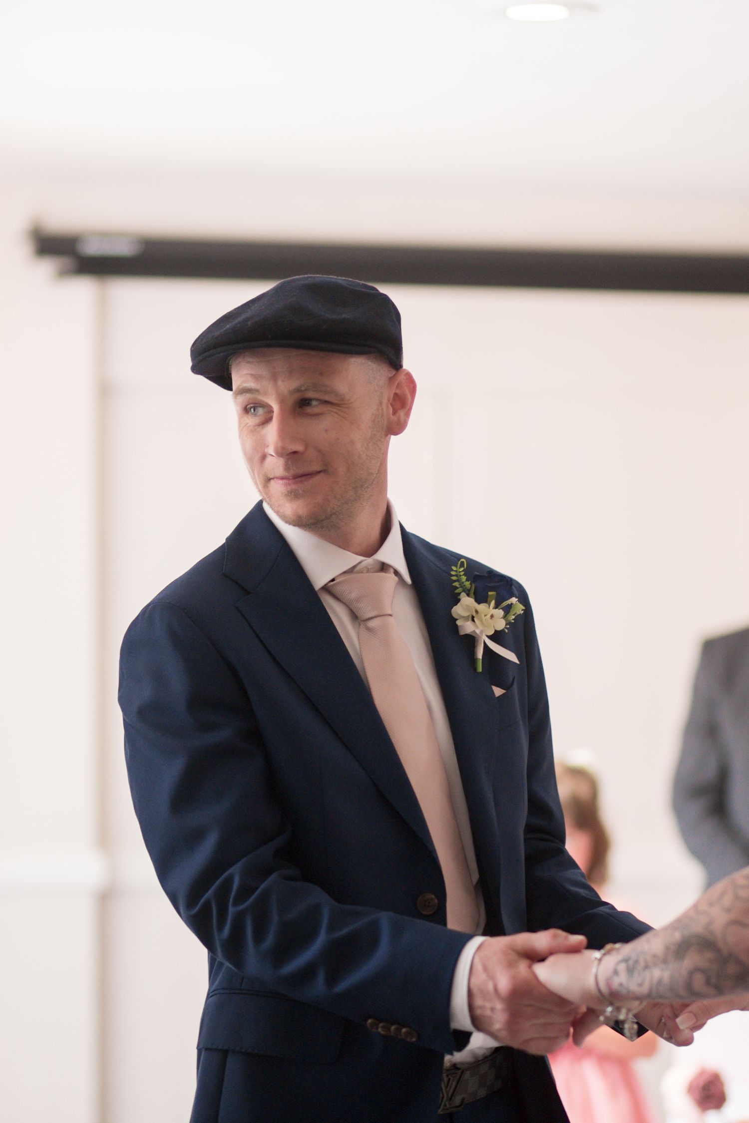 A Groom holds hands with his bride, smiling at the registrar off camera. He’s wearing a navy suit, with a light pink tie and a baker boy cap