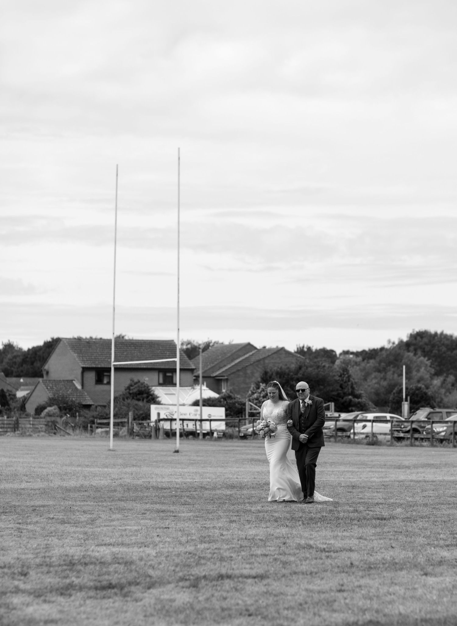 Bride and her father pass the rugby posts and cross the field on the way to the outdoor ceremony