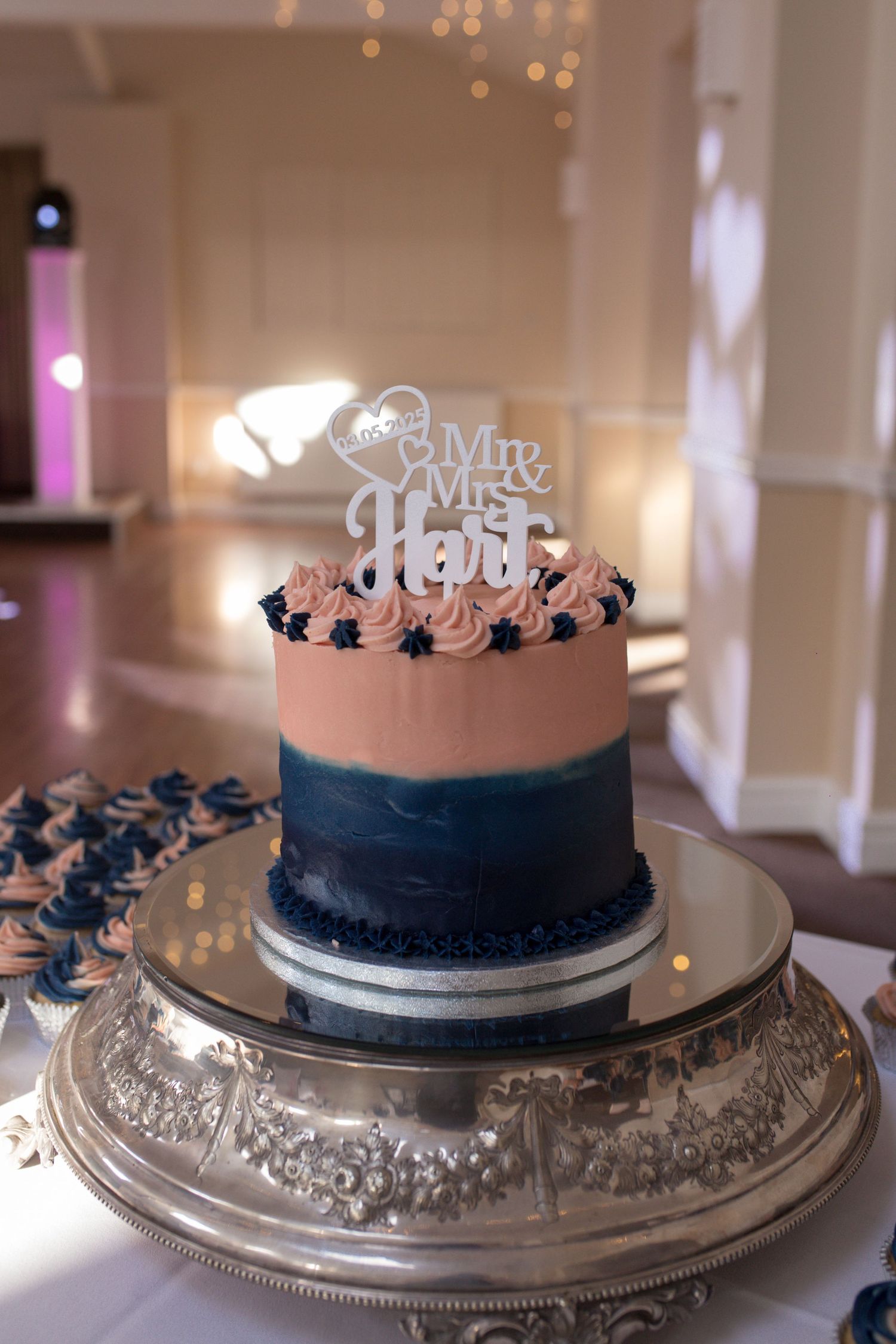 Close up of a wedding cake. Dark blue on the bottom, light pink icing on the top half, topped with a sign saying Mr & Mrs Hart