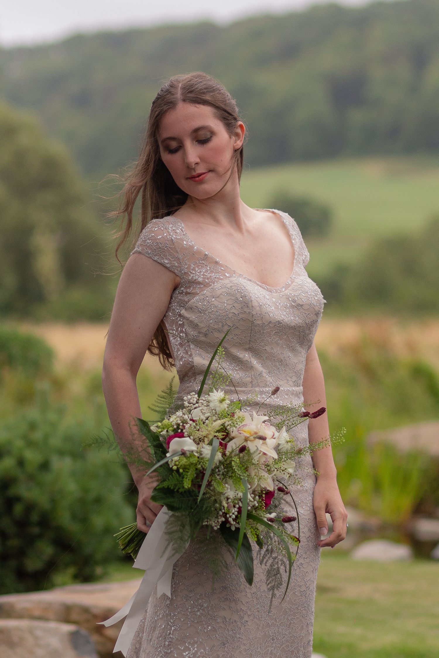 A close up of a bride wearing a silvery dress, a bouquet of flowers held at her hip and she gracefully looks down at them.