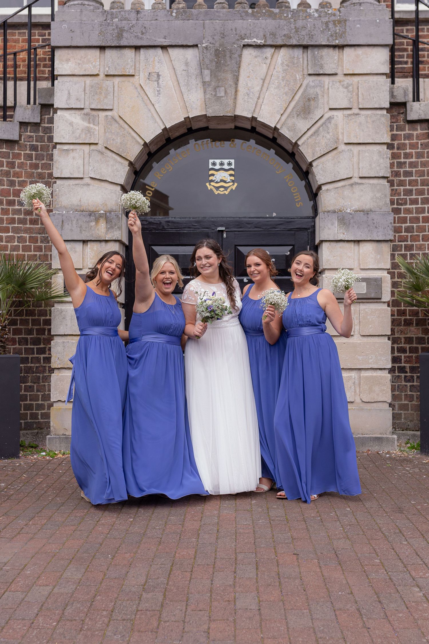 A bride and her bridesmaids celebrate during a group photo. Blue bridesmaids dresses, and bouquets held up to the sky