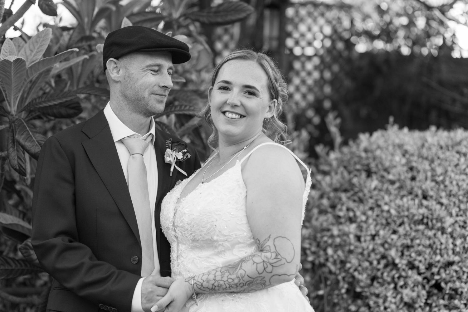 Bride and Groom pose together in the garden, bodies angled together. Bride smiles at the camera, whilst the groom looks lovingly at his wife