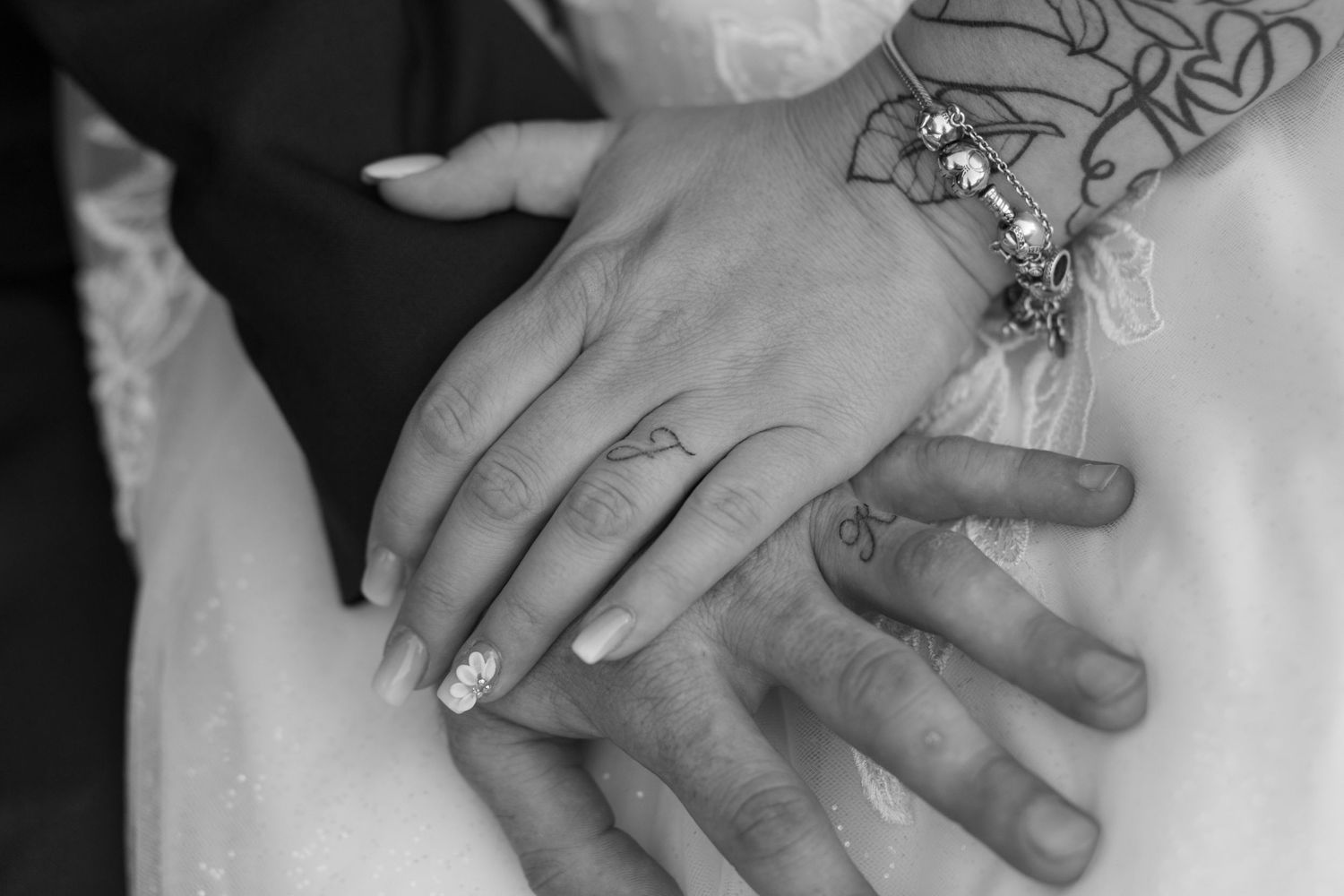Black and White, close up photo of two hands, one placed on top of the other. Tattoos of initials replace traditional wedding bands.