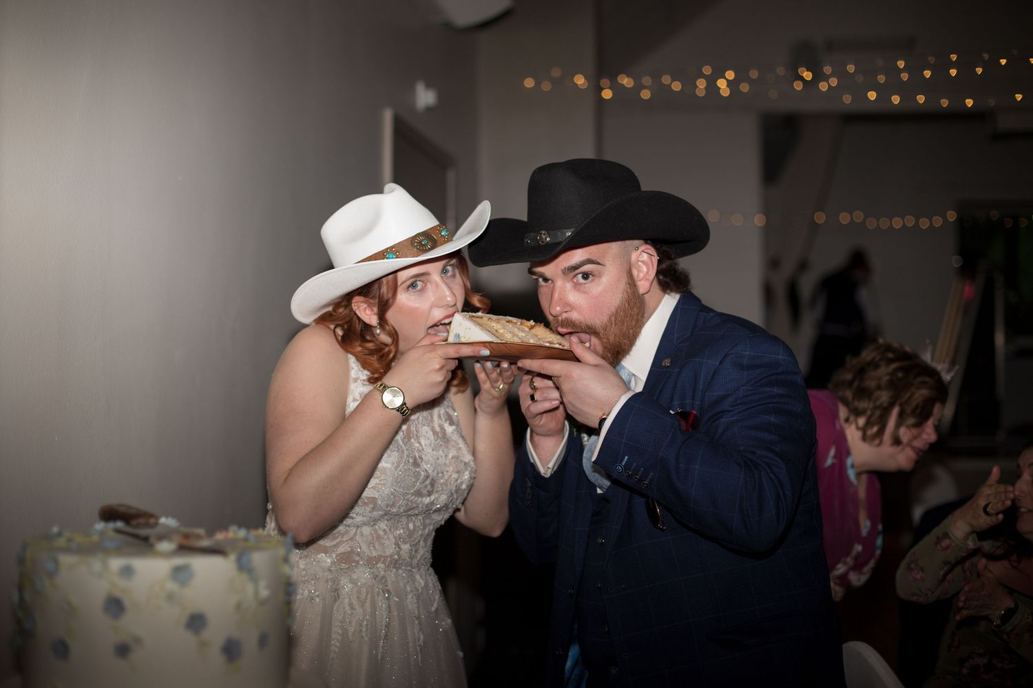 Bride and Groom both wearing cowboy hats, pose for a photo both pretending to bite into the same slice of their wedding cake.
